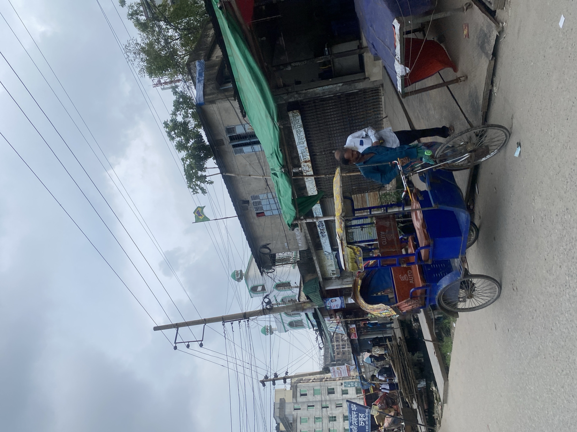 This image depicts a street scene in Dhaka, Bangladesh, on a cloudy day. In the foreground, a blue auto-rickshaw is parked on a paved road. Two men are visible near the auto-rickshaw; one, wearing a teal shirt, appears to be adjusting something on the vehicle, while the other, in a white shirt, stands beside him, looking towards the right.
The background reveals a bustling urban environment. A tall utility pole with multiple wires stretches across the frame. Buildings line the street, including a white mosque with green accents and a multi-story building with green-tinted windows. Several shops with green awnings are present, one of which has a metal grate in front. A Brazilian flag is visible flying from a building on the right. People are scattered throughout the scene, some walking and others appearing to be engaged in commerce. The overall impression is one of a busy street market or thoroughfare.