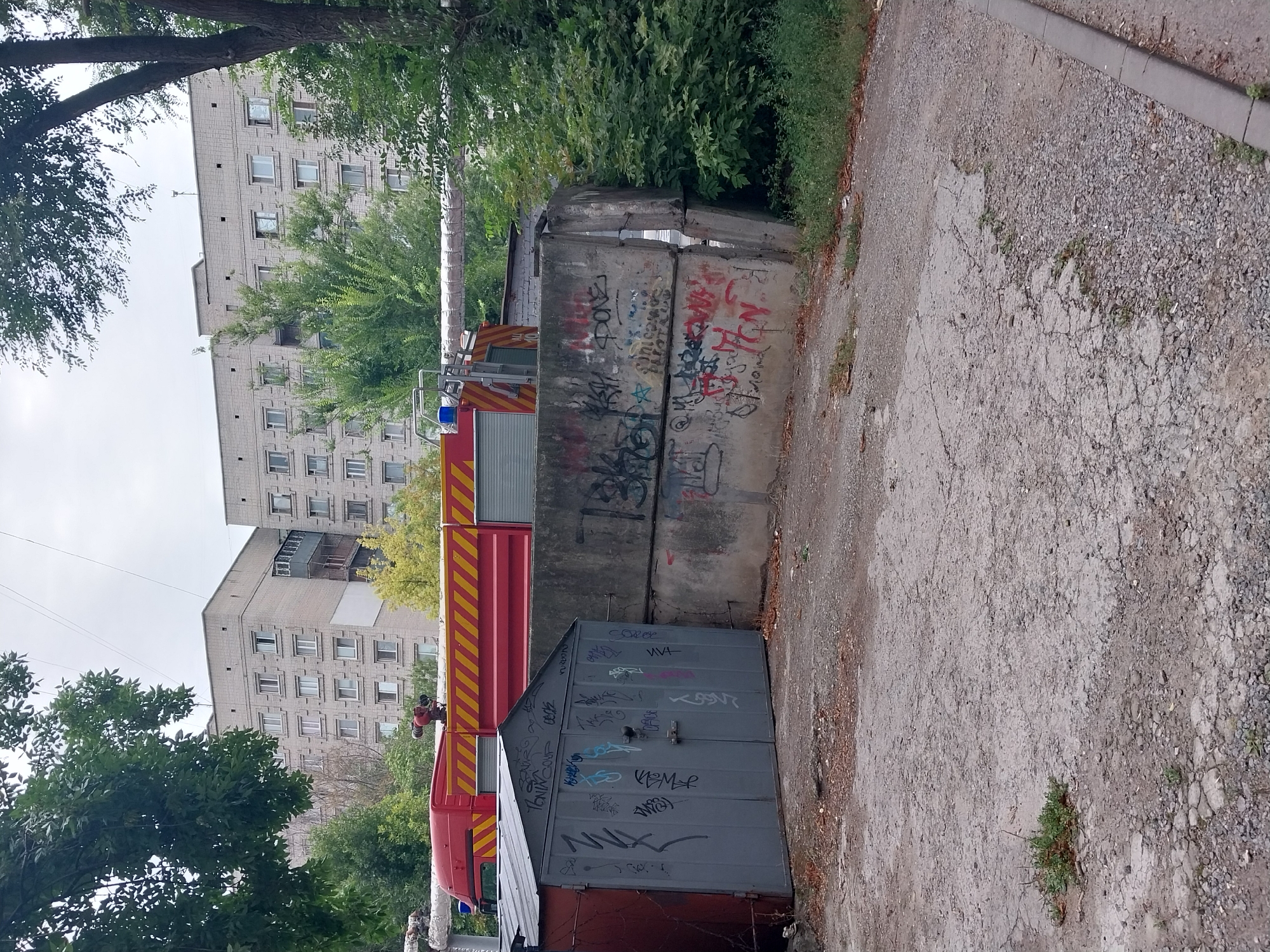 A red fire truck with yellow diagonal stripes on its side is parked behind a grey metal garage door covered in graffiti. The fire truck has a ladder mounted on top and a blue flashing light. The garage is situated in front of a concrete wall, also covered in graffiti. In the background, there are tall, multi-story apartment buildings. Trees and green foliage are visible throughout the scene, with power lines stretching across the sky. The ground is a mix of gravel and cracked pavement. This scene appears to be in an urban setting, possibly in Dnipro, Ukraine, given the architectural style of the buildings and the presence of graffiti.