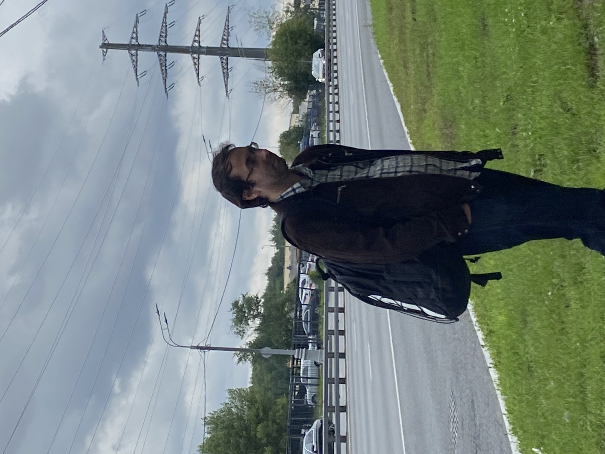 A man wearing glasses, a plaid shirt, a dark jacket, and dark pants stands on a grassy verge next to a multi-lane road. He is carrying a black backpack. The road appears to be a highway or a major thoroughfare, with vehicles visible in the background. In the distance, there is a power line pylon and trees. The sky is overcast. The scene is likely located in the city of Khimki, Russia, given the presence of power lines characteristic of the area and the general landscape.