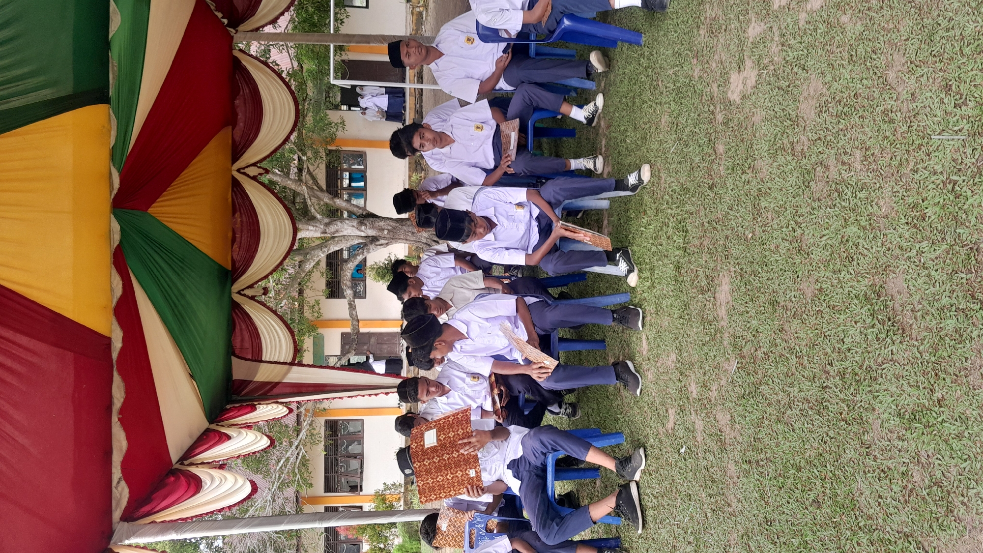 A line of young male students dressed in what appears to be school uniforms is seated on blue plastic chairs, lined up on a grassy field. Most of them are wearing black skullcaps. They are holding various objects, some with patterned covers, in their laps. The students are arranged horizontally, oriented with their left sides toward the viewer.
To the left, there is a large, multi-colored tent or canopy structure, made of fabric in alternating colors of yellow, green, red, and white. In the background, a light-colored building with windows is visible behind a tree. The location is identified as Blangriek, Indonesia.