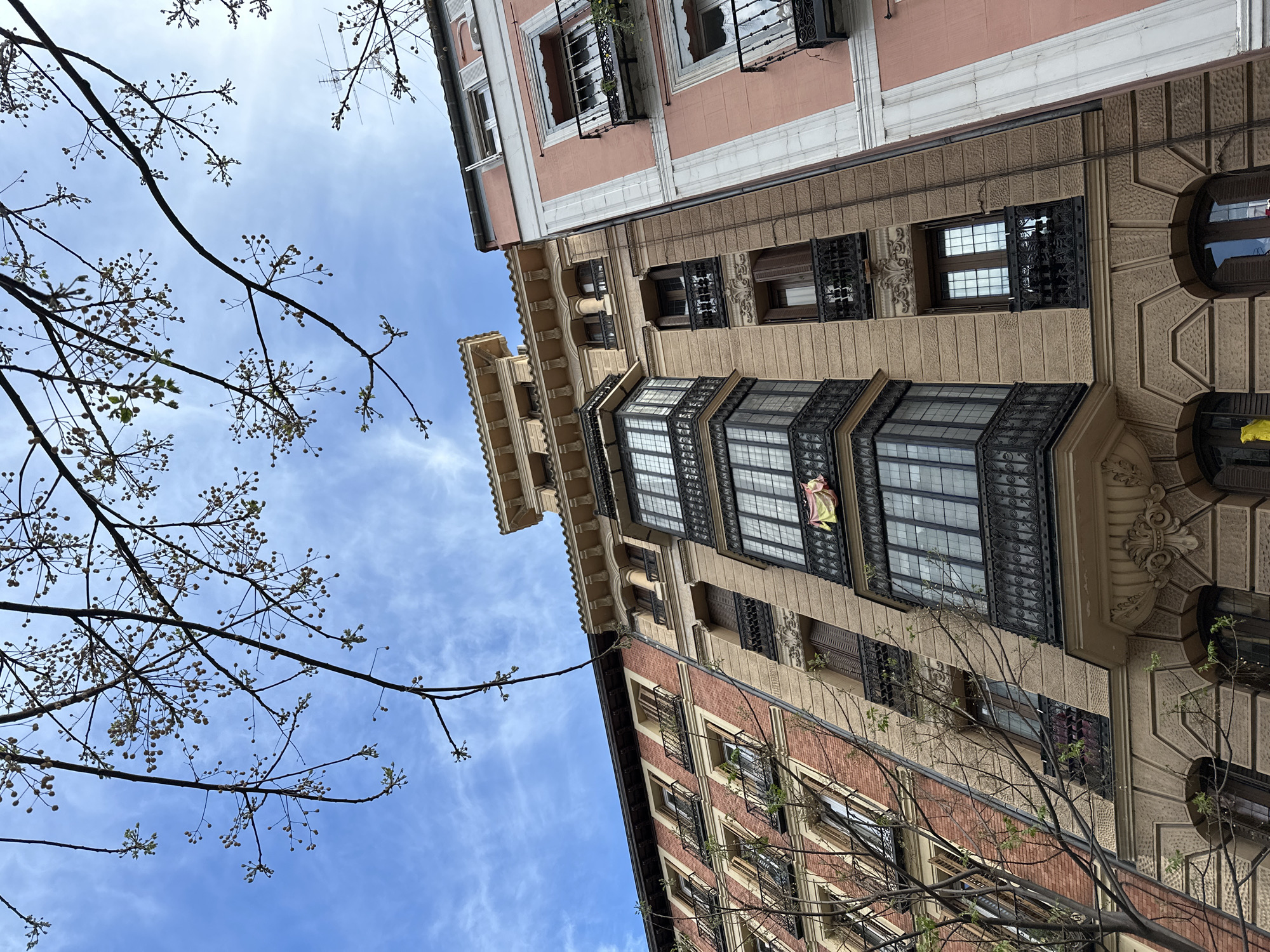 A low-angle shot shows a corner view of two buildings in Madrid, Spain, under a partly cloudy blue sky. The buildings are adjacent, one pink and the other composed of beige and red brick. The beige building has distinctive architectural features including a bay window on the upper levels with dark, ornate ironwork and several smaller windows with similar detailing. A flag is partially visible hanging from one of the windows in the bay. Parts of tree branches with budding flowers frame the buildings on the left and the right sides of the picture.