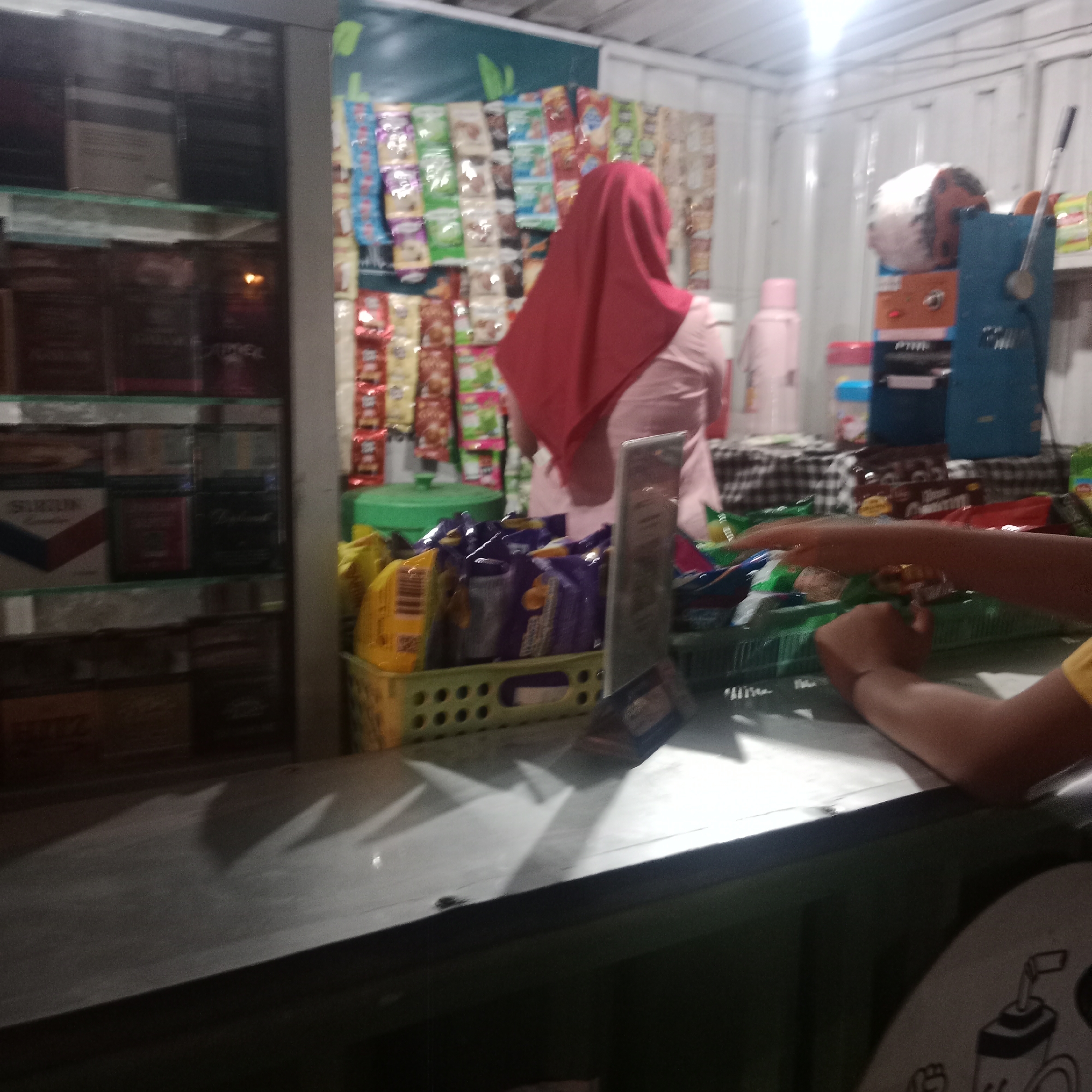 The image depicts a small, indoor stall, likely a convenience store or warung, in Tegowanu Kulon, Indonesia. The stall is filled with various products, predominantly snacks and beverages, displayed on shelves and in baskets. In the background, a woman wearing a red hijab and a light pink shirt is visible, appearing to be attending to the stall. In the foreground, a person's arms are visible, suggesting they are interacting with the products or the counter of the stall. A counter or table runs across the lower portion of the image, with several baskets of packaged snacks placed upon it. The lighting suggests it is either evening or the stall has artificial lighting. The walls appear to be corrugated metal, common in such structures in Indonesia. The overall impression is of a small, bustling local business.