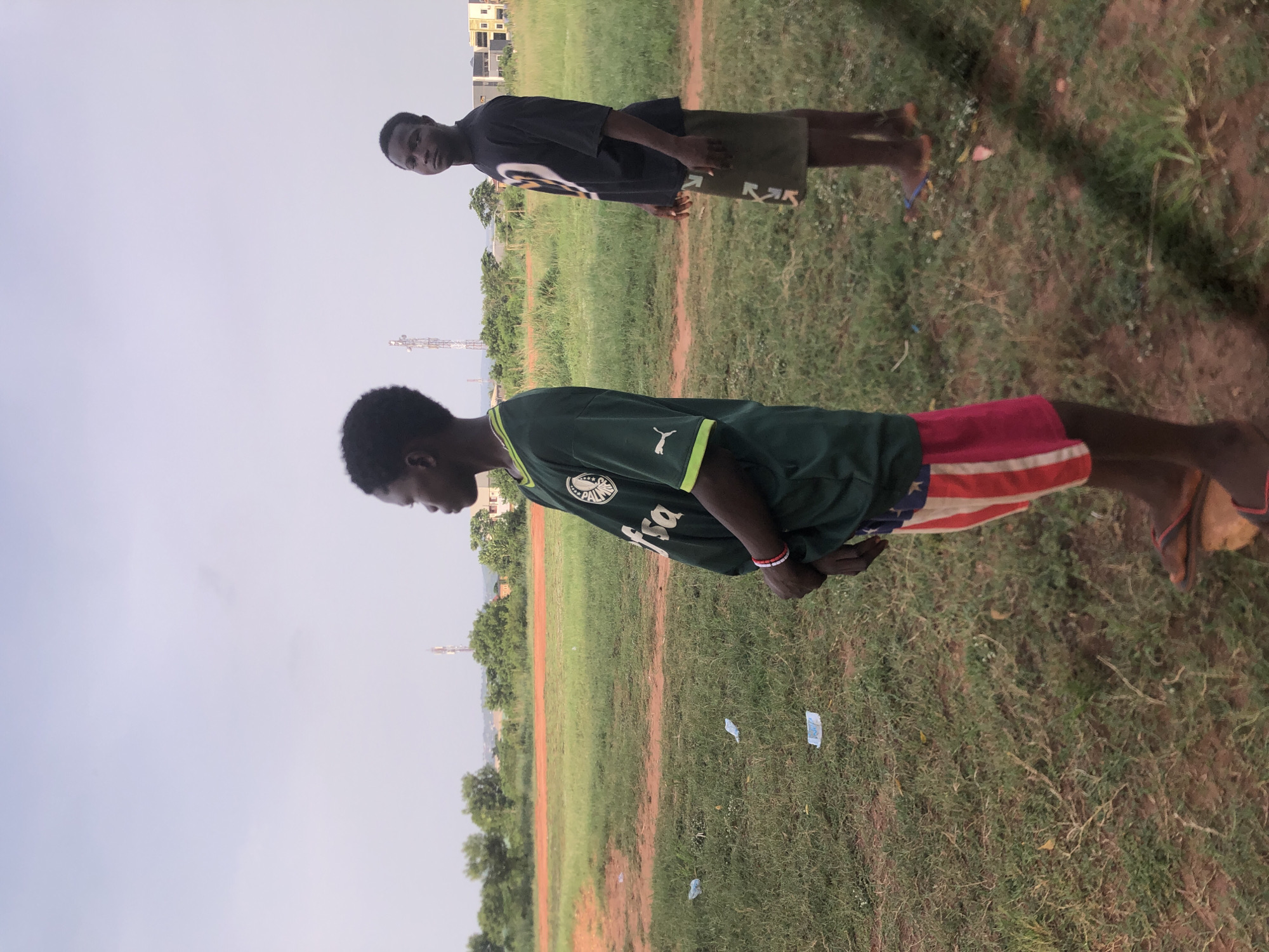 The image displays two young males standing in an open, grassy field under a bright, overcast sky, characteristic of a daytime scene in Ukwu-Abwa, Nigeria.
In the foreground, a male with dark skin and short dark hair is visible, oriented to the left. He is wearing a dark green short-sleeved football jersey emblazoned with the "PALMEIRAS" logo, a white "Puma" logo, and "Psa" text. His shorts are distinctively patterned in red, white, and blue, reminiscent of the American flag. A multi-colored bracelet (red, white, black) adorns his left wrist, and he appears to be barefoot.
Further back and to the right, another young male with dark skin and short dark hair stands, facing and looking towards the viewer. He wears a dark-colored (possibly black or dark grey) short-sleeved t-shirt with a prominent white graphic on the front, likely a brand logo (resembling "OFF"). His shorts are dark green or brown with white design elements. He is also barefoot, with blue flip-flops visible on the ground next to his foot.
The setting is an expansive field with patchy green grass and visible dirt paths. In the background, there are trees, sparse vegetation, and several light-colored residential buildings with dark roofs. A tall communication tower is also visible on the horizon. A few small pieces of white litter are scattered on the grass. The overall activity appears to be casual standing or resting in an outdoor environment.