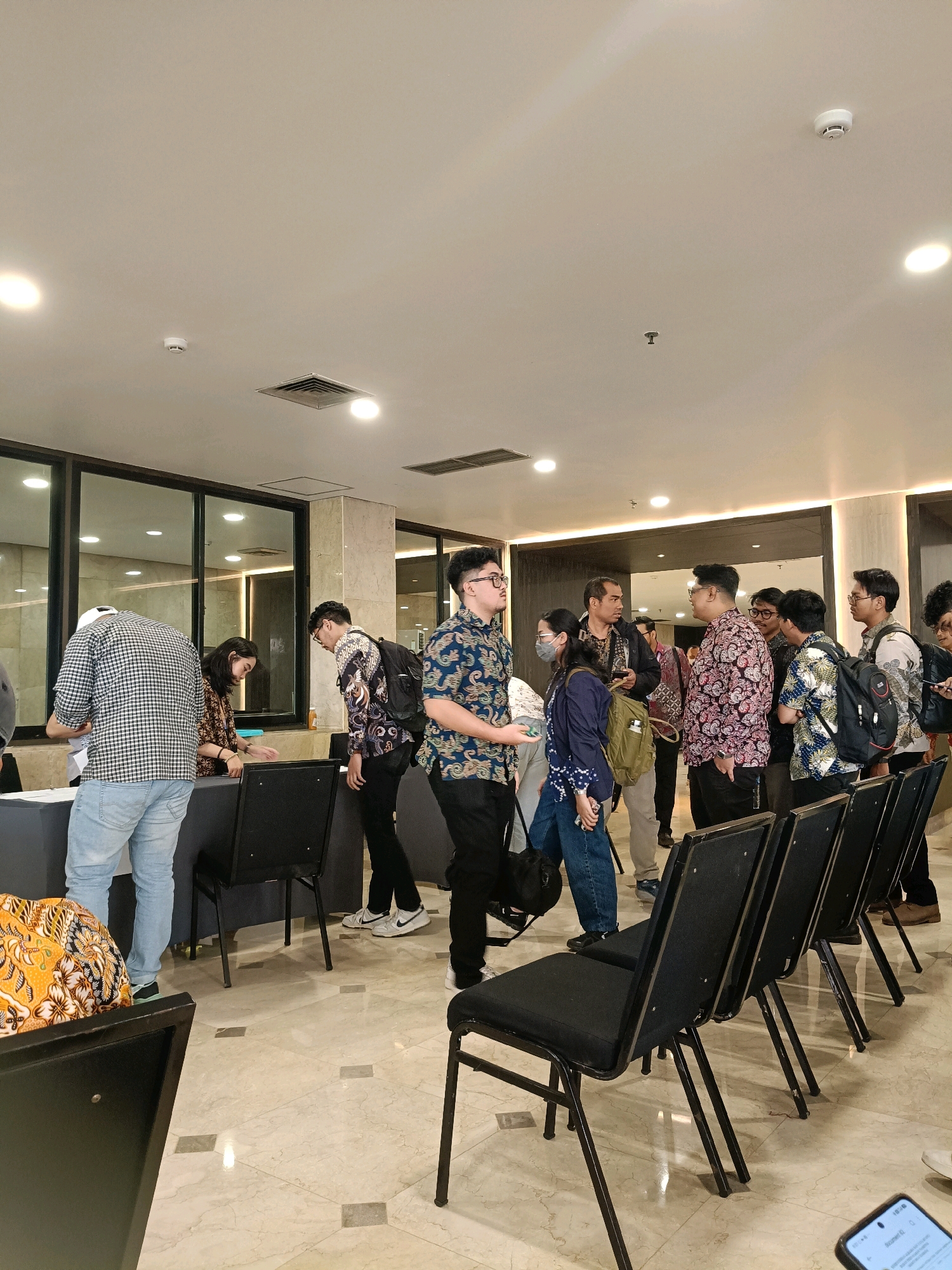 The image depicts an indoor scene, possibly a convention or registration area, with a group of people standing. The environment features a marble-tiled floor, black chairs arranged in rows, and a long black table in the foreground where some individuals are interacting with what appears to be registration materials. The walls have a polished, marble-like finish and are illuminated by recessed lighting.
In the foreground, to the left, a person wearing a black and white checkered shirt and light blue jeans stands facing away from the camera, looking down at the table. Next to them, a woman with dark hair is leaning over the table. To their right, a man in a batik shirt with a black backpack is also engaged at the table. Further to the right, a group of men are standing and conversing. They are mostly wearing batik shirts, suggesting a cultural event or conference. One man in a dark blue batik shirt is speaking to another man in a pink and black batik shirt. A woman with a mask and a dark blue top is standing between them. The lighting is bright and even, typical of an indoor public space. The overall impression is of an organized gathering or event. The location is specified as a city in North Jakarta, Indonesia.