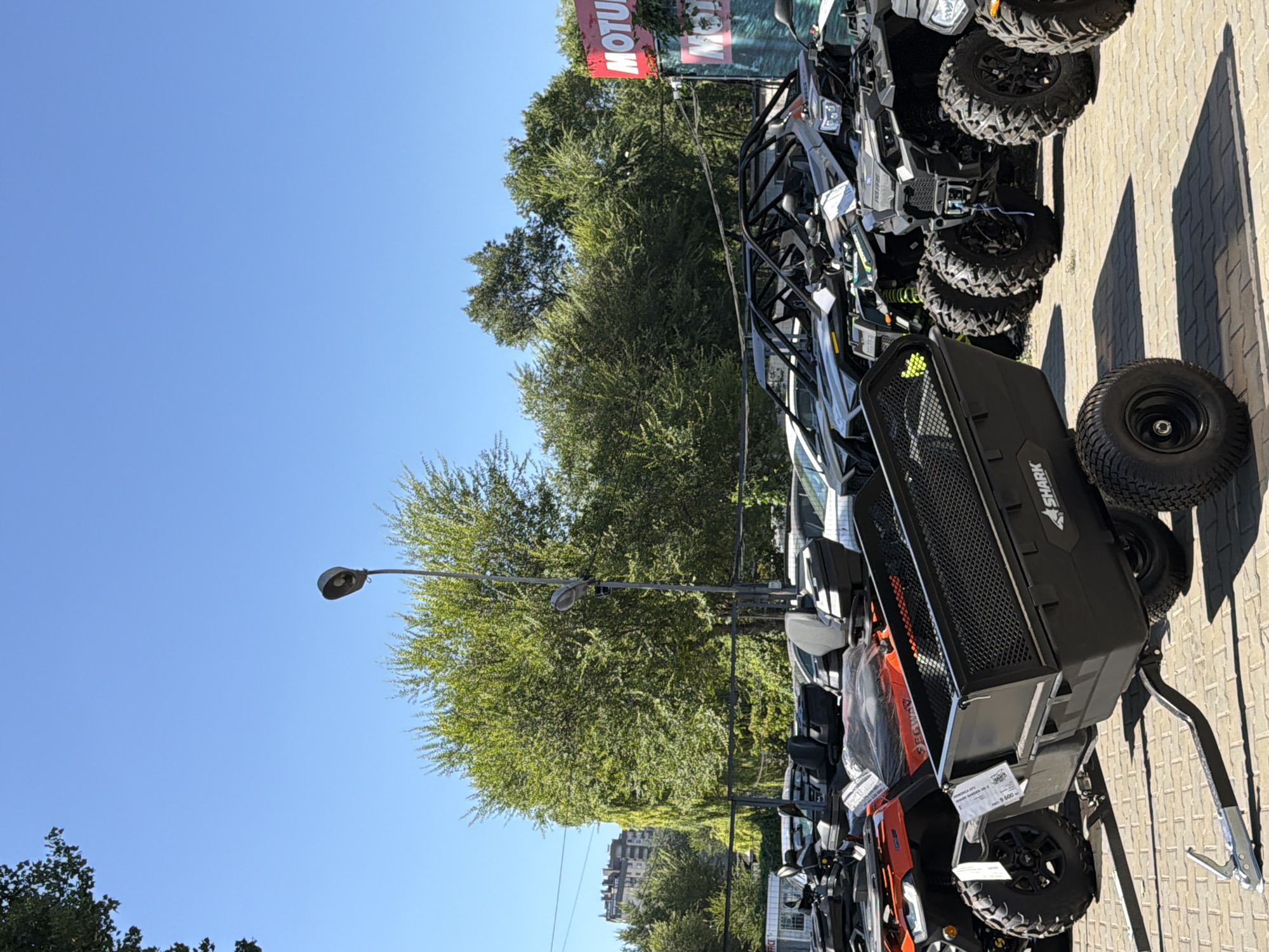 The image depicts an outdoor scene, likely a parking lot or showroom, featuring several all-terrain vehicles (ATVs) and a small trailer. In the foreground, a black trailer with a mesh top is hitched to an unseen vehicle. To the left of the trailer is an orange ATV, and behind it, more ATVs and what appear to be off-road buggies are parked in rows. In the background, lush green trees provide a natural backdrop against a clear blue sky. A tall lamppost is also visible amongst the trees. A red and white sign with the word "MOTUL" prominently displayed is situated on the right side of the frame, suggesting the location might be a dealership or service center. The ground is paved with light-colored bricks. The overall impression is of a sunny day at an outdoor recreational vehicle establishment.
While the image itself does not contain explicit markers of its location in Chișinău, Moldova, the presence of the "MOTUL" sign is a common indicator of automotive businesses, and such establishments are found in cities worldwide. The architecture visible in the far background, a multi-story building, could be characteristic of urban environments. Without further contextual clues within the image, pinpointing the exact street or specific area within Chișinău is not possible. However, the scene strongly suggests a commercial area geared towards vehicles and outdoor recreation.