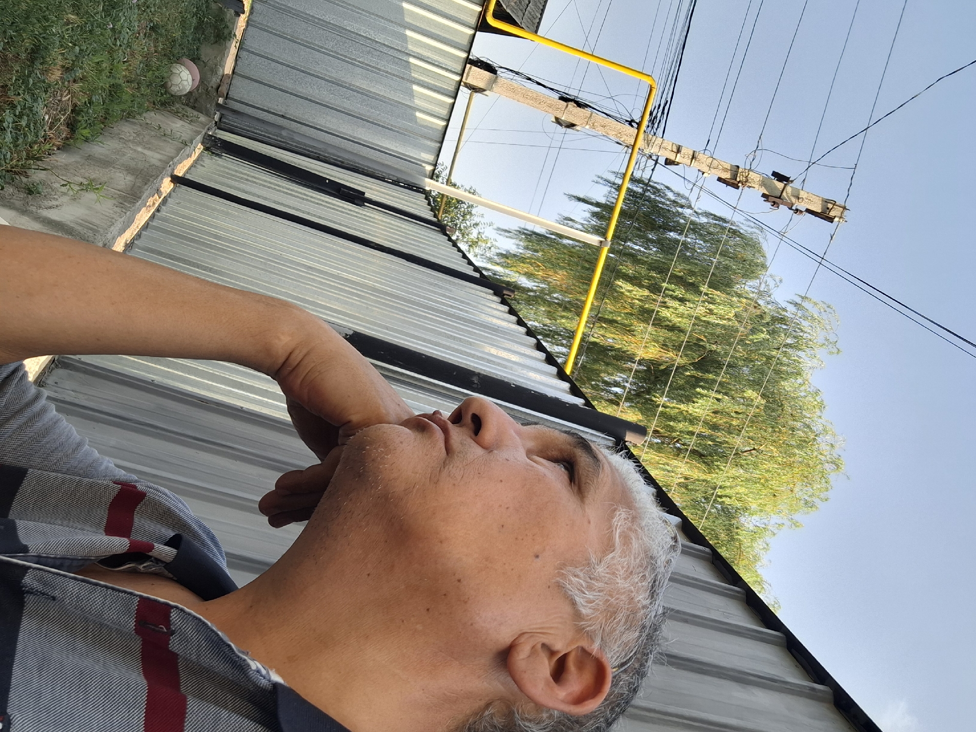 An adult male with graying hair is shown in a close-up, profile view, resting his chin on his hand. He is looking to the left, away from the camera, with a thoughtful or pensive expression. He is wearing a horizontally striped polo shirt with red and navy blue accents. The background is composed of a corrugated metal fence, a concrete border, and some greenery. In the distance, there is a utility pole with power lines and a yellow gas pipe, along with a weeping willow tree. A partially visible soccer ball is on the grass near the concrete border. The sky is clear and blue, suggesting it is daytime. The overall impression is an outdoor scene, possibly a residential yard or a utilitarian area within the city of Almaty, Kazakhstan, characterized by industrial and natural elements.
