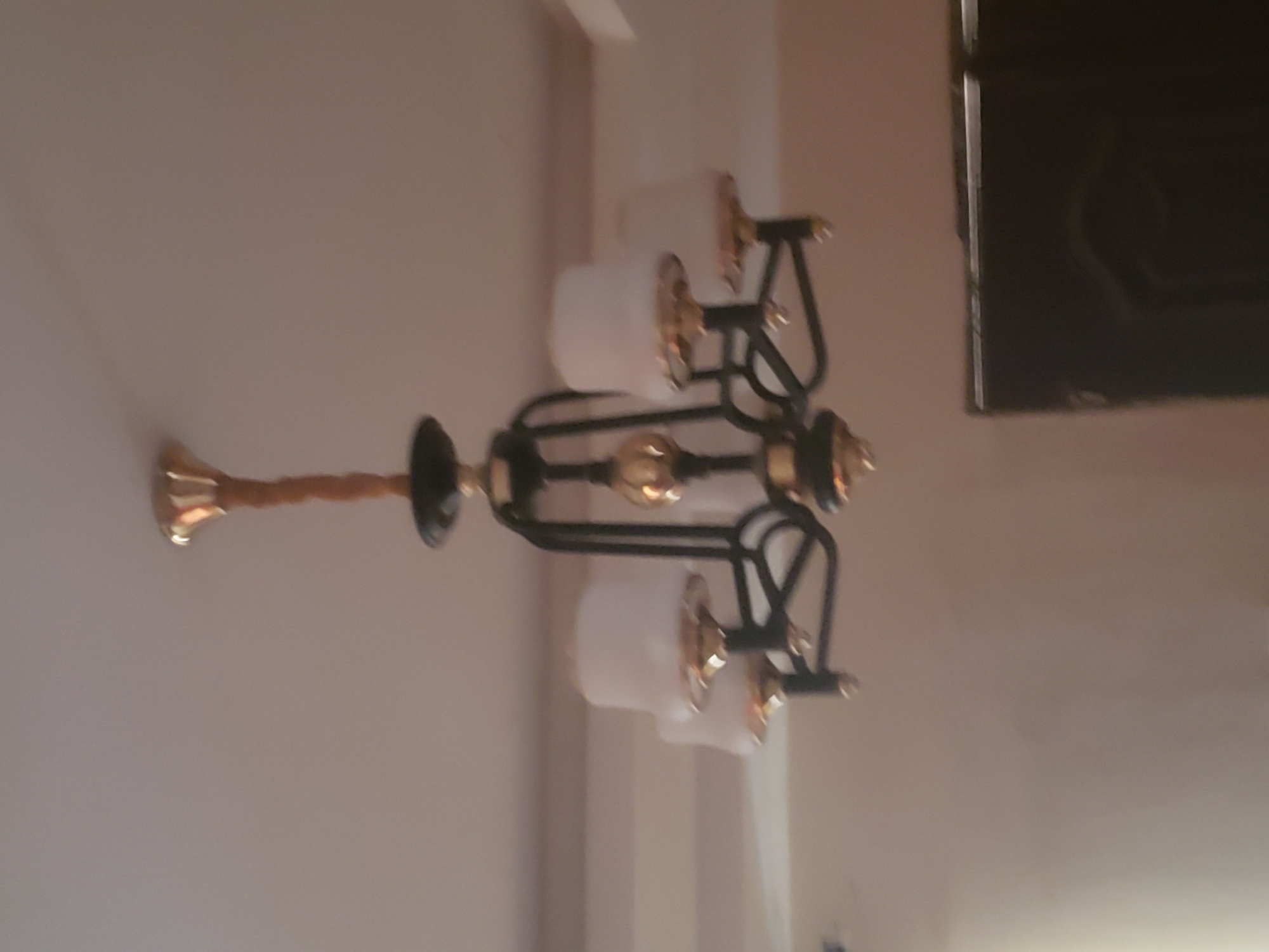 A chandelier hangs from a ceiling in a room. The chandelier has a central gold-colored stem wrapped in a brown material, from which black metal arms extend. Each arm supports a white lampshade with gold trim. The lampshades appear to be in the shape of upside-down cups. The ceiling is a neutral color. In the background, a dark, indistinct object, possibly a door or furniture, is visible. The image is blurry and lacks sharp detail. There are no people or actions depicted.
