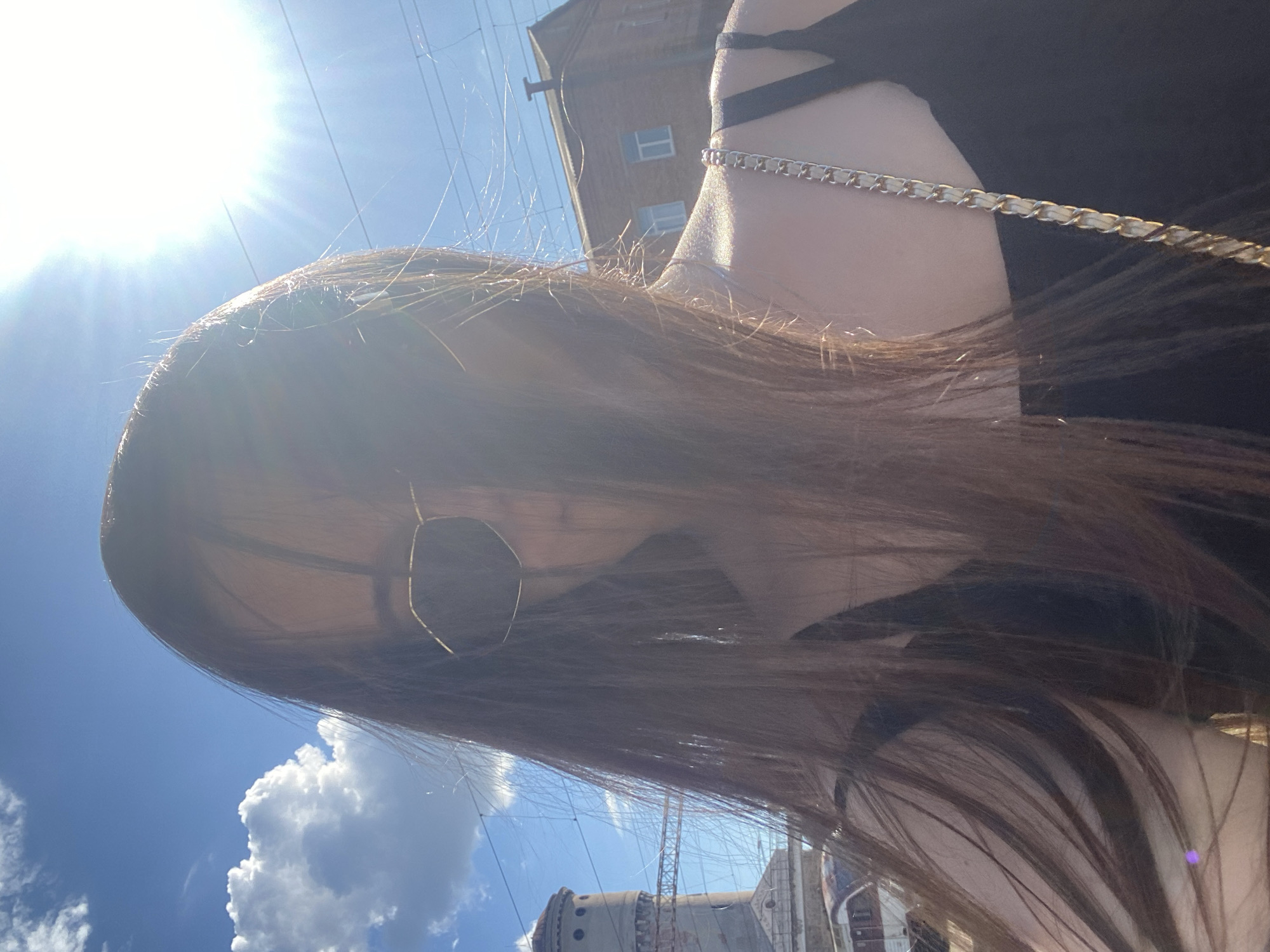 The image shows a young woman with long, brown hair, wearing sunglasses and a black top with a gold chain strap. She is taking a selfie outdoors on a sunny day. The background features a blue sky with scattered white clouds, a tall brick building with windows, and some electrical wires overhead. To the left, there is a distinctive cylindrical tower with circular windows, which is a known landmark in Poltava, Ukraine. The overall scene suggests a casual outdoor setting, possibly near a train station or industrial area, given the visible infrastructure. The sun is bright and high in the sky, casting a lens flare across the image.