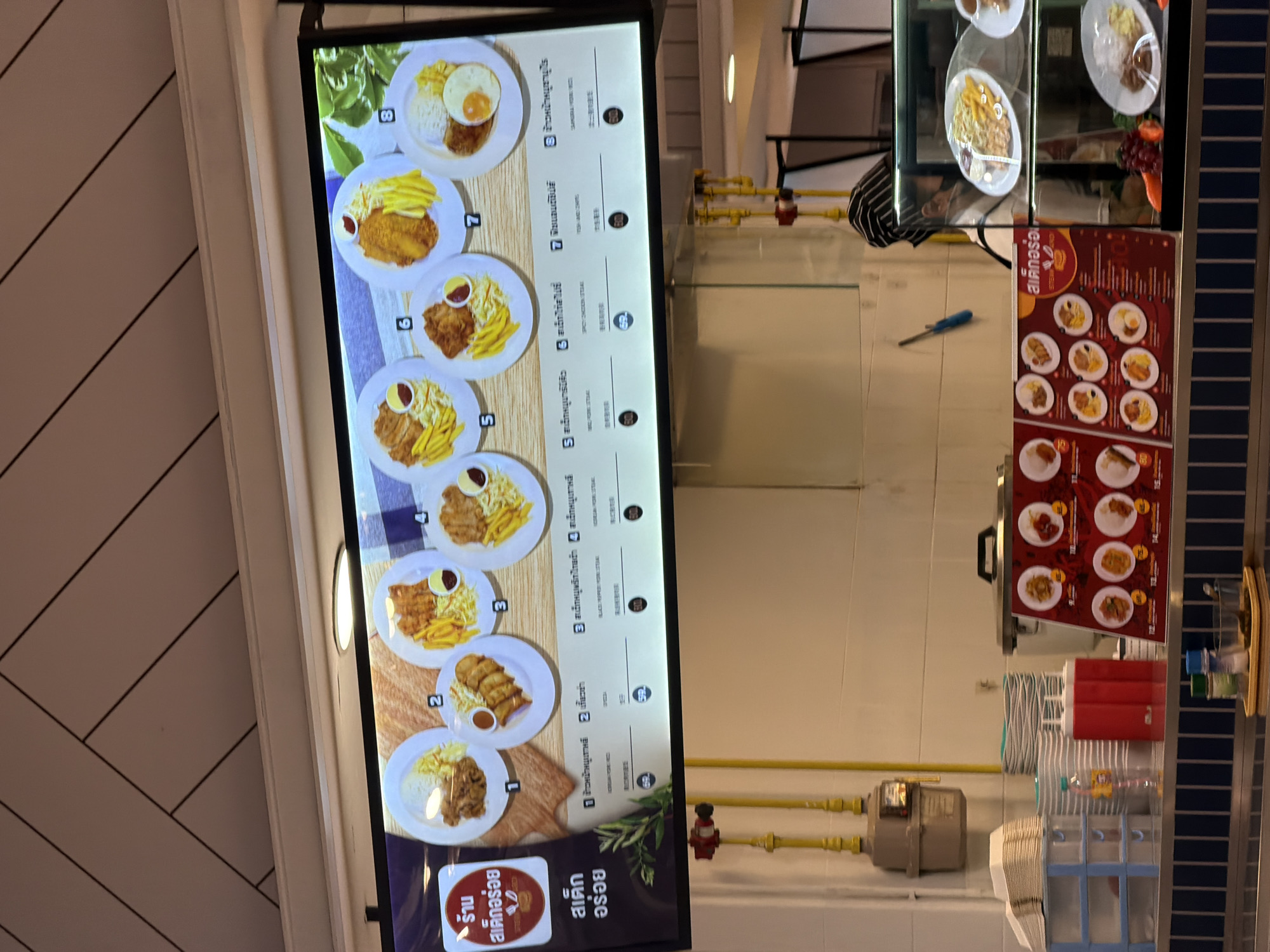 The image shows a digital menu board displaying various food items, likely in a restaurant or food court. The menu is in Thai and English, with items numbered from 1 to 8. The food items appear to be mostly meat dishes served with fries and rice. Below the digital menu, there is a red poster board with smaller circular images of food items, also with prices. To the left of the menu, there is a stack of disposable plates and cups. Further to the left, there is a gas meter attached to the wall. To the right of the digital menu, a glass display case shows some of the dishes available, and a person wearing a chef's hat and mask is visible behind it, suggesting food preparation is ongoing. The overall environment appears to be indoors, possibly a food establishment in Baan Bang Rak, Thailand.