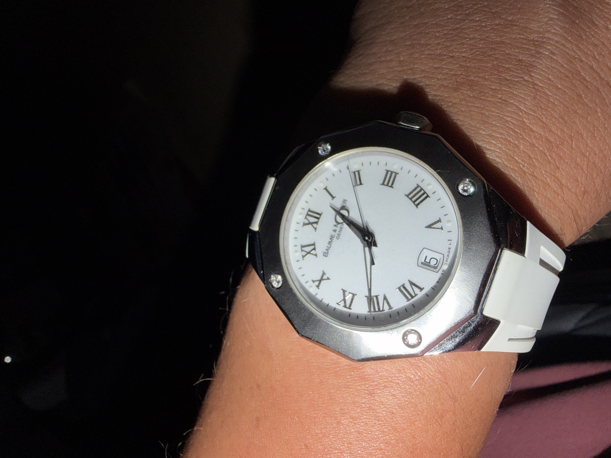 A close-up, eye-level shot shows a Baume & Mercier watch on a person's wrist. The watch has a white dial with Roman numerals and a date window showing "5". The watch case is octagonal and silver-toned, with four diamond hour markers. The strap appears to be white silicone. The background is dark and out of focus. No specific location in Haifa, Israel, can be identified from the image.