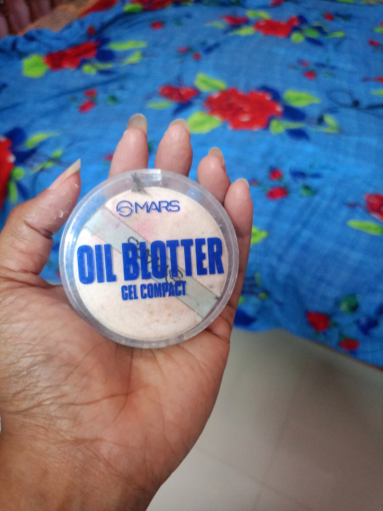 A person's hand, with visible palm lines and fingernails, holds a round, clear plastic compact containing a light-colored pressed powder. The compact lid features a blue logo that reads "MARS" and text that says "OIL BLOTTER" in large blue letters, with "GEL COMPACT" in smaller blue letters below. A diagonal band of light blue and pink stripes is visible across the powder within the compact. The background is a blue fabric with a red and green floral pattern. The image was taken in Chenkavila, India.