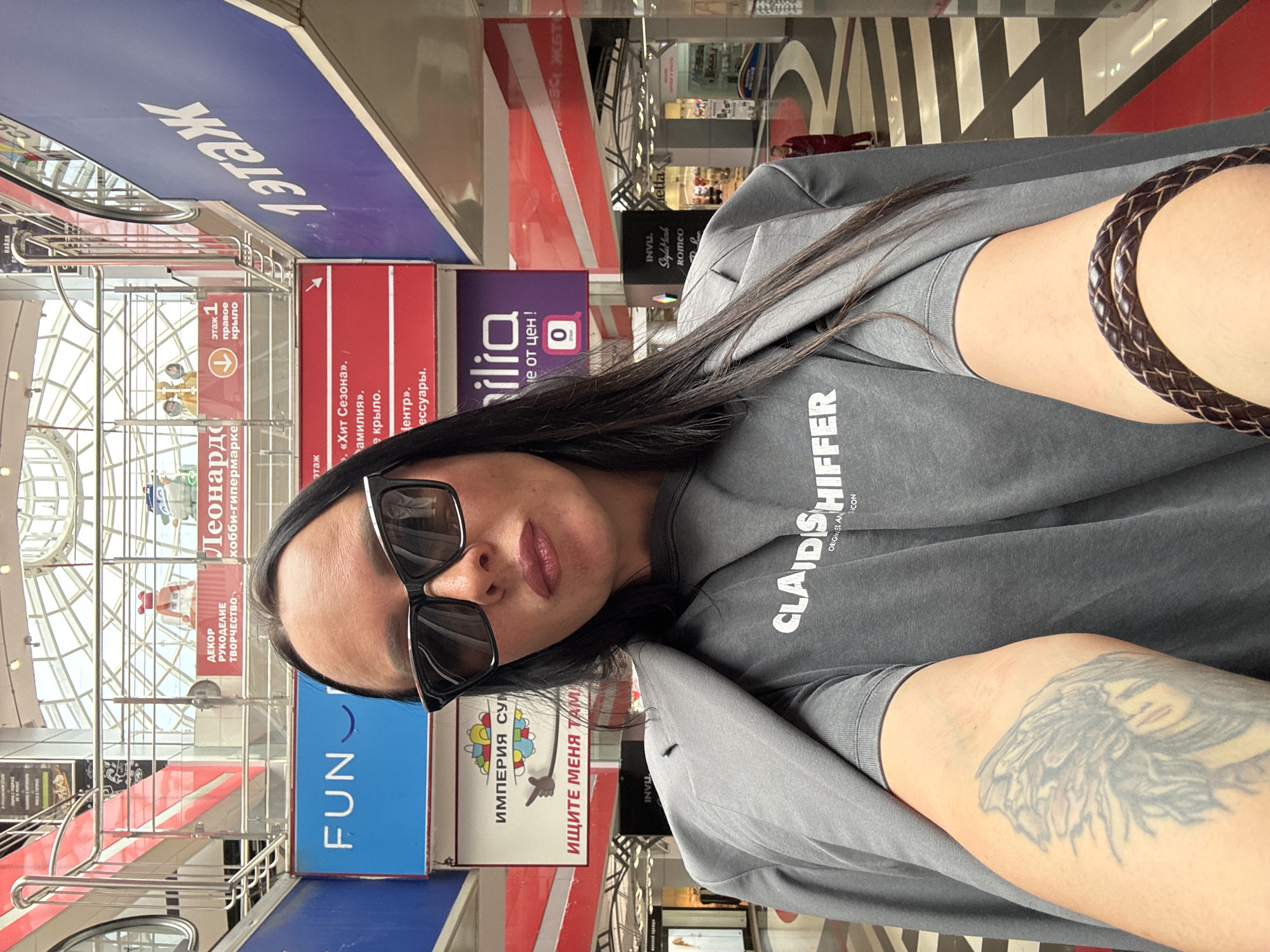 A medium-shot image shows a person wearing sunglasses and a gray sweatshirt with the word "CLAID SHIFFER" printed on it. The person has long, dark hair and a brown braided bracelet on their left arm, which also has a tattoo. The location appears to be a multi-story shopping mall in Izhevsk, Russia, based on the presence of signage in Russian. The backdrop features store signs for "Леонардо," "FUN," "ИМПЕРИЯ СУМ," and "ilia," as well as navigational signs indicating directions. The mall has a structured ceiling and a decorative floor with black and white patterns. The perspective is slightly angled.