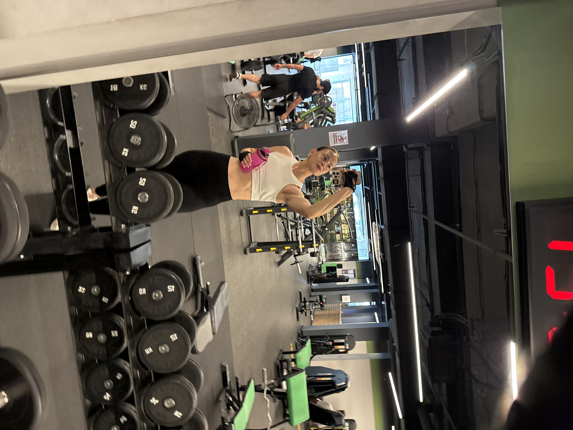 A woman is taking a mirror selfie in a gym. She is wearing a white sports bra and black leggings, and holding a pink water bottle. In the background, other gym-goers are visible using equipment, including a man on a treadmill and another person on a weight machine. The gym appears to be well-equipped with various weights, machines, and mirrors. The overall environment suggests a typical indoor fitness facility.