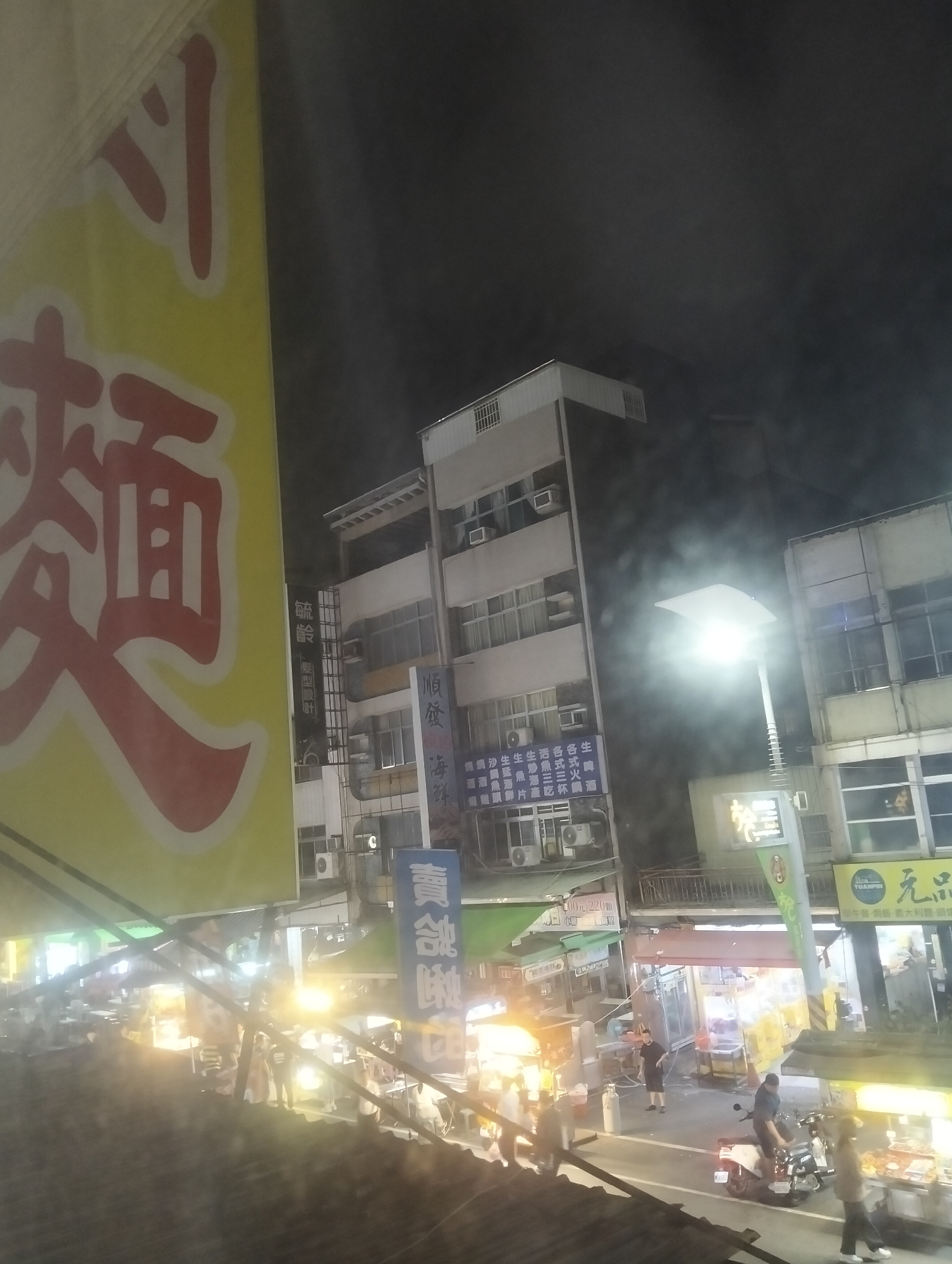 The image depicts a night scene in a street market, likely in Kaohsiung, Taiwan, given the presence of Chinese characters and the typical urban night market atmosphere. In the foreground, a large yellow banner with red Chinese characters is visible, partially obscuring the view of the street. The characters on the banner appear to be related to food, possibly noodles based on the visible characters.
The street itself is bustling with activity. Numerous food stalls are set up along the street, illuminated by bright lights, creating a vibrant and energetic ambiance. People are seen walking around, interacting with vendors, and purchasing food. Some individuals are operating the stalls, preparing food, or attending to customers. A few motorcycles are parked on the side of the street.
The background features multi-story buildings, typical of an urban environment. These buildings have illuminated windows and signage, indicating shops or residences. A tall street lamp with a bright, modern design is prominently featured in the mid-ground, casting a strong light on the street and surrounding area. The sky is dark, suggesting it is nighttime. The overall impression is a lively and active night market, a common feature of Taiwanese cities.
