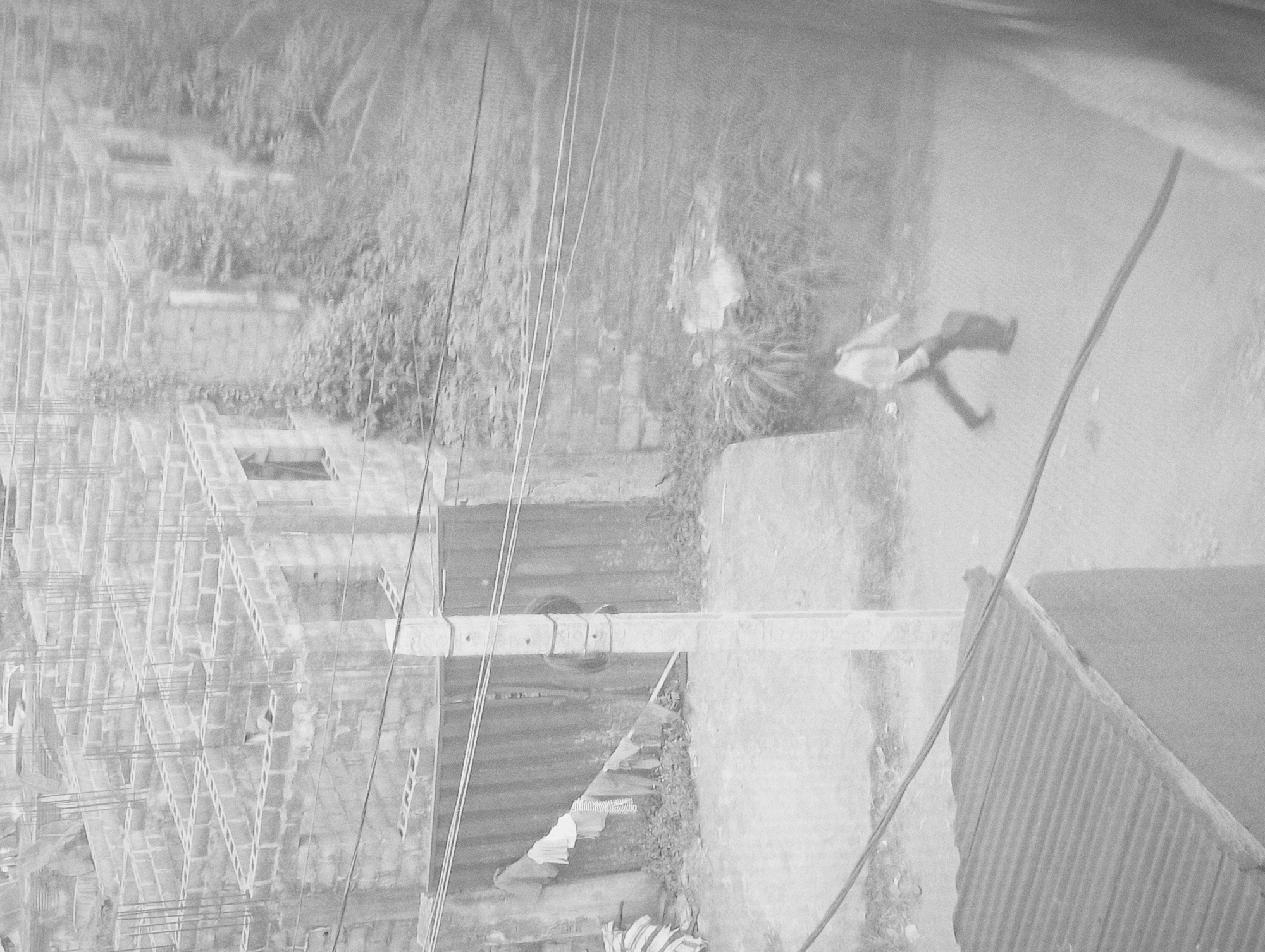 The image, taken in Nkpor, Nigeria, presents a street scene from an elevated perspective. A man wearing light-colored clothing walks along a concrete walkway next to a road. The environment is characterized by an unfinished building constructed of concrete blocks and steel supports. Overhead, multiple electrical wires run along wooden poles. An old tire hangs on one of the poles. A corrugated metal roof is visible in the bottom right corner of the image. Vegetation is seen in the background. Garments are placed above some sheet metal.