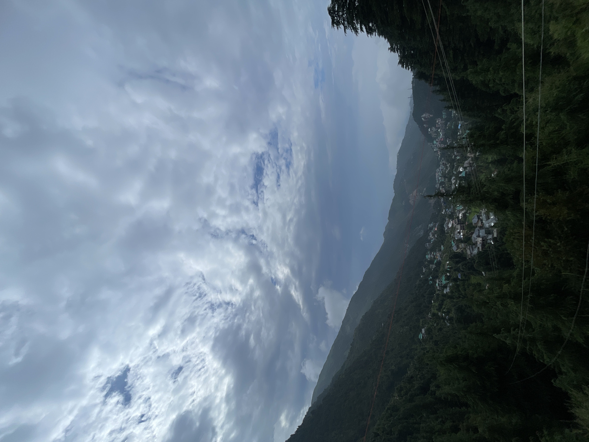 The image captures a panoramic view of a mountainous landscape with a village nestled within the valley, likely in Haini, India. The foreground is dominated by dense, dark green coniferous trees, with the tips of some reaching into the frame. Several thin, dark wires are stretched horizontally across the lower portion of the image, suggesting the presence of electrical or communication lines. Beyond the trees, the slopes of the mountains are covered in vegetation, and a cluster of buildings, representing the village, is visible in the middle ground. The buildings have diverse roof colors, including green, grey, and red. The terrain is steep, with the village appearing to cling to the mountainside. In the background, more mountains recede into the distance, shrouded in a misty haze. The sky is overcast with dramatic clouds, ranging from light grey to dark, with patches of blue sky peeking through in the upper left quadrant. The overall atmosphere is one of natural beauty and serene, albeit cloudy, weather. There are no visible people or animals in the image.