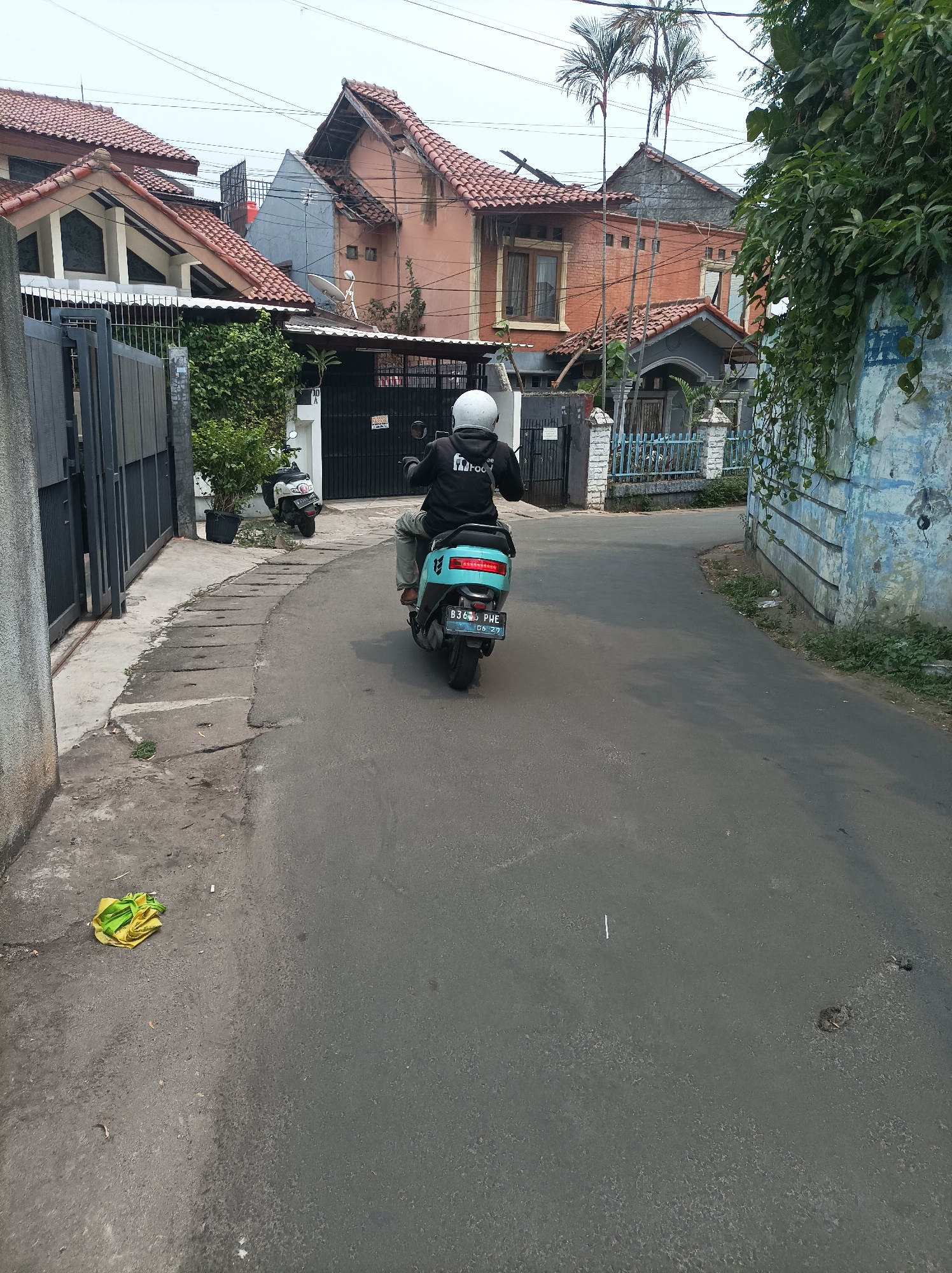 In West Jakarta, Indonesia, a person wearing a white helmet and a black hoodie with white lettering is riding a teal electric scooter down a paved road. The scooter has a license plate that reads "B 3465 PHE". The rider is facing away from the camera. The road is bordered on the left by a sidewalk with a concrete drain and a metal fence. A yellow and green crumpled plastic bag lies on the sidewalk. On the right side of the road, there is a blue and white painted wall covered in vines. Behind the wall, houses with red tile roofs and brick or painted facades are visible. Two other scooters are parked near a black gate on the left side of the road, further up. The sky is overcast.
