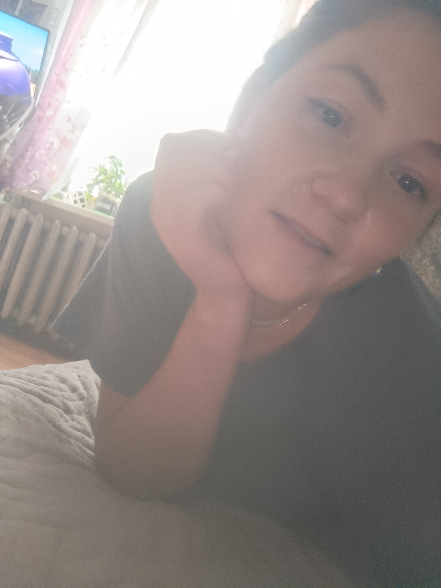 The image is a close-up, over-the-shoulder selfie taken indoors, likely in a residential setting. The subject, a woman with light brown hair, is lying down and resting her head on her hand, looking towards the camera. She is wearing a dark grey t-shirt and a delicate necklace. The background shows a window with sheer, patterned curtains, through which bright sunlight is streaming in, causing lens flare. To the left of the window, a television screen is visible, displaying a colorful image. Below the window, a white radiator runs horizontally across the wall. The woman is positioned on a textured grey blanket or bedspread. The image does not contain any elements that would indicate a specific location in the city of Karsha, Russia.