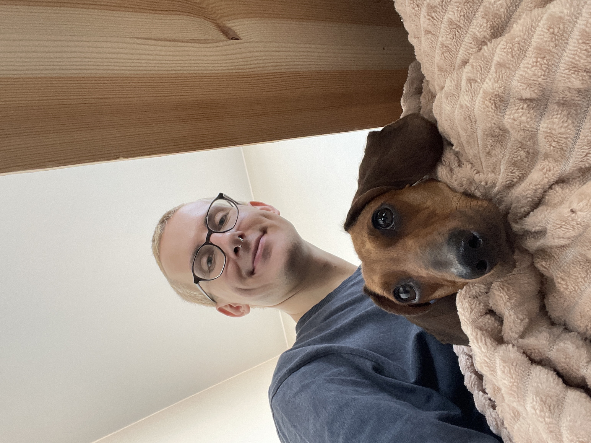 In the foreground of the image, a dachshund's face is visible, with its head resting on a beige, textured blanket. The dog has brown fur, dark eyes, and floppy ears. Behind the dog, a person with short blonde hair, glasses, and a nose ring is smiling. They are wearing a dark blue t-shirt and are partially visible, with their head tilted downwards. The background is a mix of a plain white ceiling and a wooden structure, possibly part of a ceiling or wall. The lighting suggests an indoor setting. No specific location in Saint Petersburg, Russia, can be determined from the visual information provided.