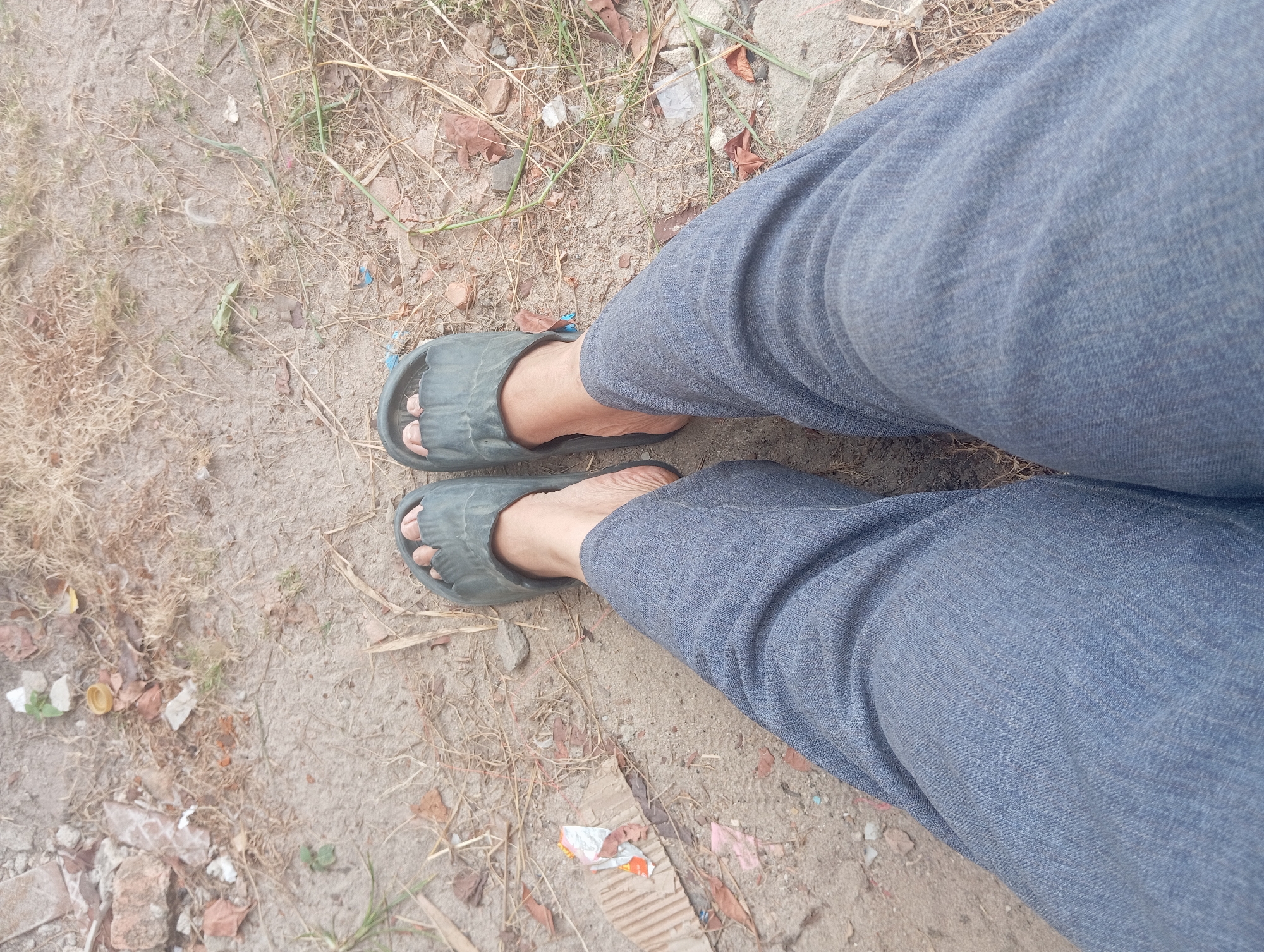 The image shows a close-up, top-down view of a person's legs wearing blue denim jeans and dark grey slide sandals, standing on a dirt and dry grass surface. Debris, including broken bricks, dry leaves, and some unidentifiable white and orange plastic fragments, are scattered across the ground. The ground is uneven and appears dry. The scene does not provide any specific visual cues to identify the exact location within Dente Teladas, Indonesia, other than the general description of an outdoor, somewhat unkempt area.