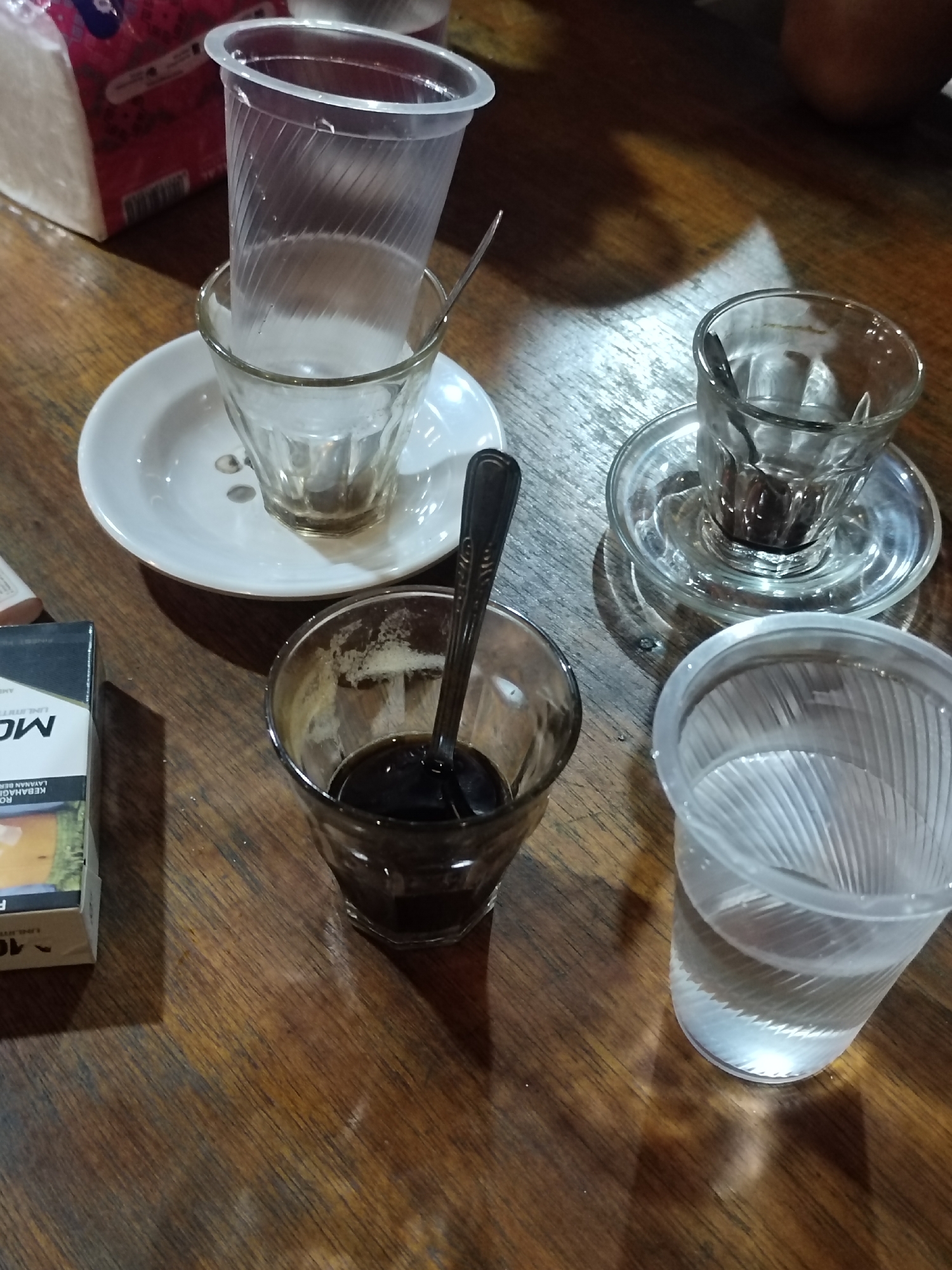 This is a close-up, eye-level shot of a wooden table with several glasses and a cigarette pack. The scene appears to be set in an informal establishment, possibly a cafe or warung, in Keudematangkuli, Indonesia.
In the foreground, a glass contains dark liquid, likely coffee, with a spoon resting inside. To the right, another glass is filled with clear water. Behind these, a small white saucer holds a clear glass with a dark residue at the bottom, possibly from a previous drink. Stacked on top of this glass is a clear plastic cup, empty. To the left of the coffee glass, a pack of cigarettes is visible, with partial text including "MO" and "10" along with Indonesian text that seems to be a warning about health.
The lighting suggests it could be daytime, with shadows cast on the table, indicating diffused light. The wooden table has a worn texture, adding to the casual atmosphere. The overall mood is relaxed and everyday, implying a moment of pause or a casual meeting. The presence of coffee and cigarettes suggests a common break or social activity. No people are visible in the frame.