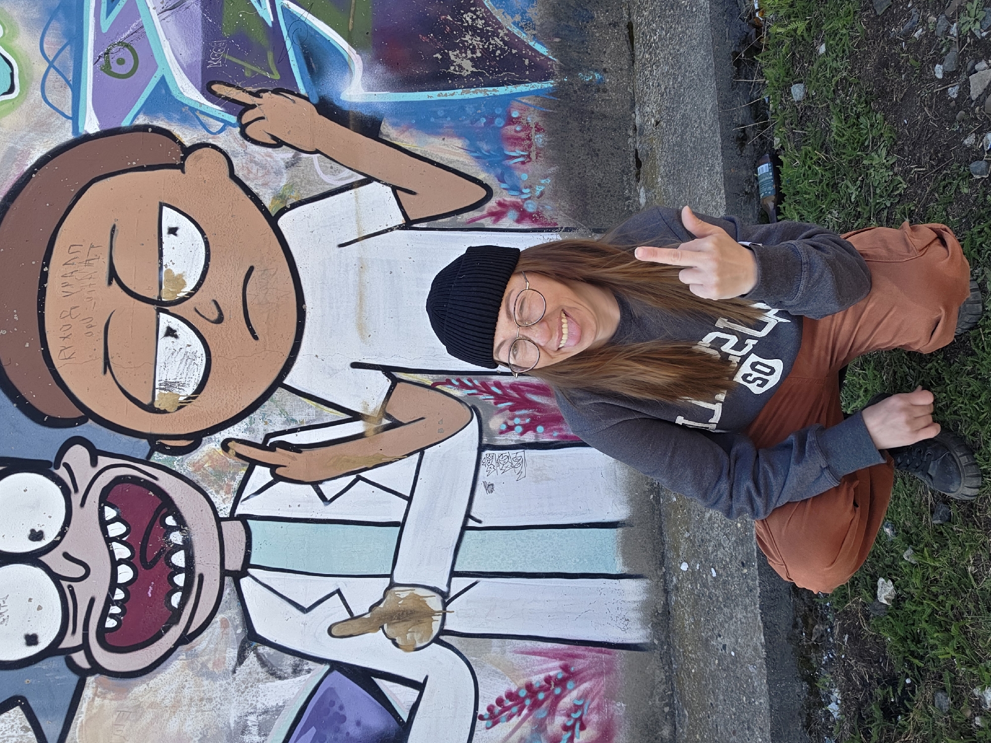 A woman wearing a black beanie, glasses, a gray sweatshirt, and orange pants is crouching next to a graffiti wall in Chaikovsky, Russia. The graffiti features cartoon characters, including one resembling Rick Sanchez from "Rick and Morty". The woman is sticking out her tongue and giving the middle finger with both hands. The ground is a mixture of concrete and grass.