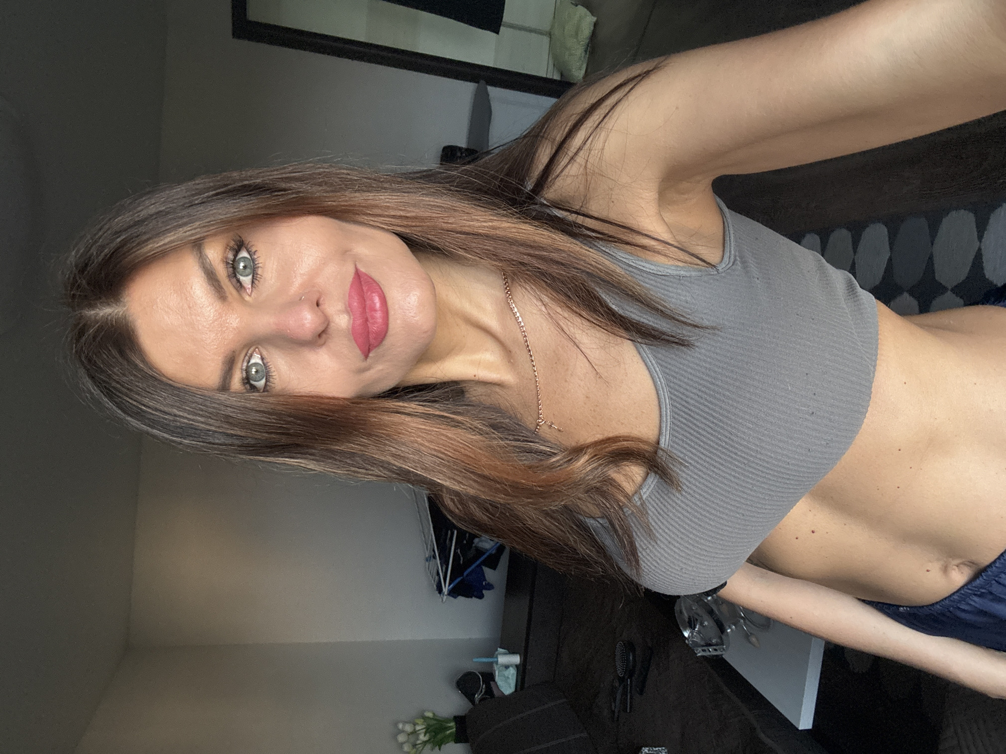 A woman is posing for a selfie indoors. She has long brown hair, blue eyes, and pink lipstick. She is wearing a grey crop top and a necklace. The location is possibly in Chaikovsky, Russia. The background includes a mirror, a cabinet, and a bouquet of white flowers.