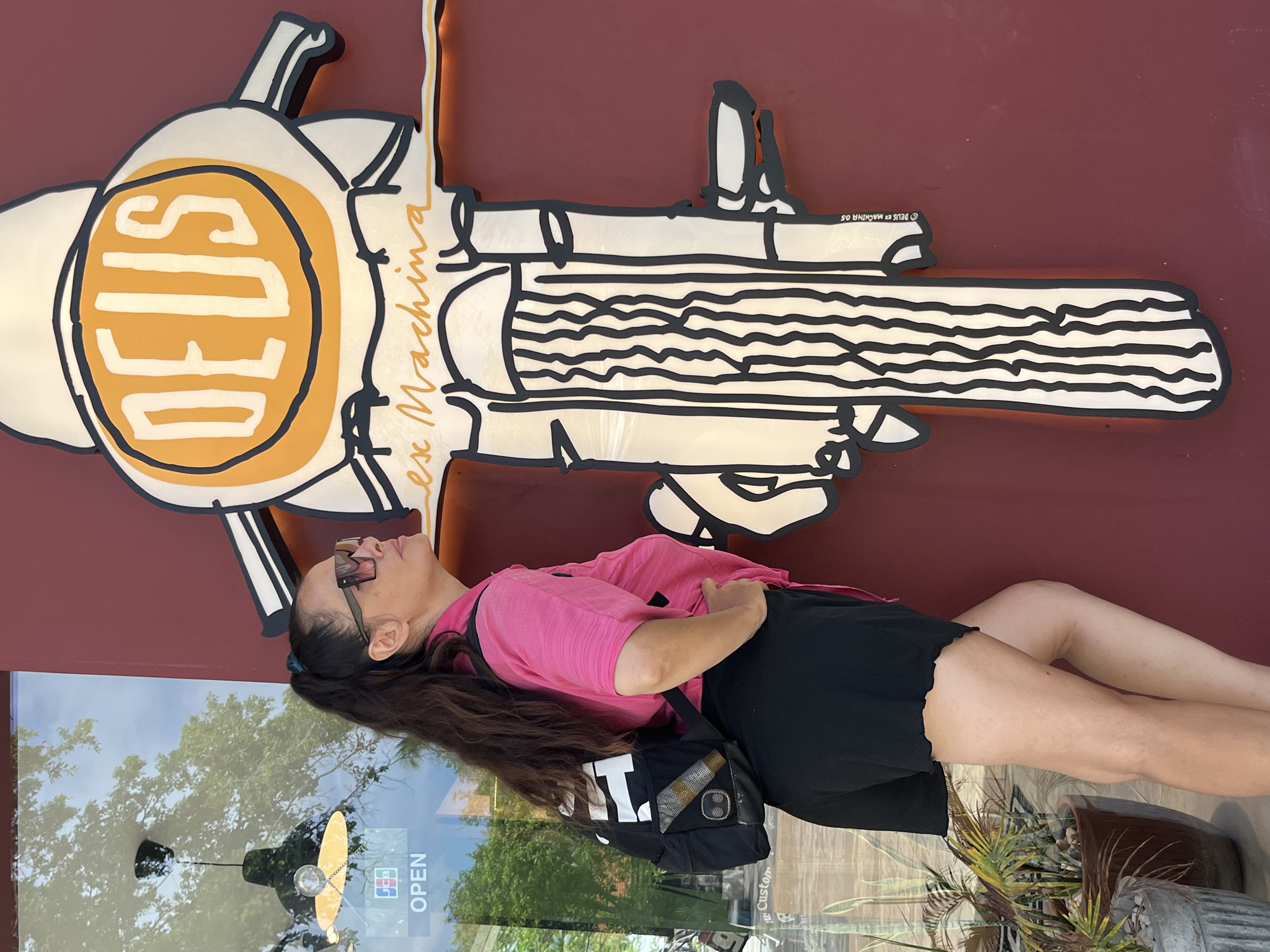 A woman with dark hair pulled into a ponytail, wearing oversized pink sunglasses, a bright pink collared shirt, and black shorts, stands with her back to the camera, facing a large, stylized graphic. The graphic is a cartoonish representation of a motorcycle front, with the word "DEUS" emblazoned in white on an orange circle at the top, and "ex Machina" written in script below it. The motorcycle graphic is mounted on a dark red wall, which appears to be an exterior wall of a building. To the left of the woman, a glass window reflects trees and a hanging light fixture. A sign with the word "OPEN" is visible on the window. The woman has one hand on her hip and is looking up at the graphic. She is wearing a black backpack with white and grey accents. The scene is outdoors, likely on a street or sidewalk, given the presence of plants in pots visible in the lower left corner. The overall setting suggests a commercial establishment in Kuta Selatan, Indonesia, potentially a surf shop or cafe, given the brand "Deus Ex Machina" which is known for its association with surf and motorcycle culture.