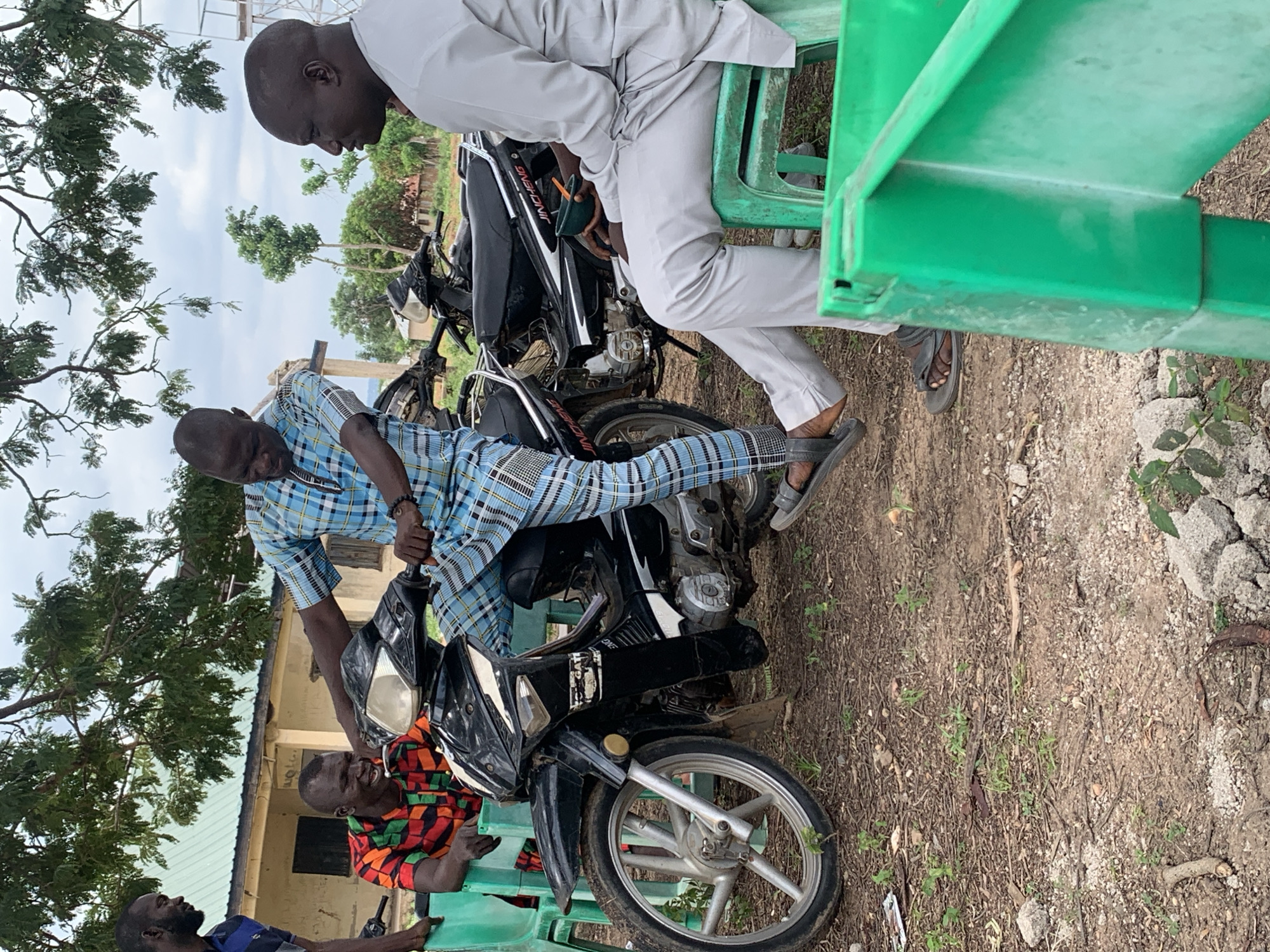 In Akwanga, Nigeria, three adult males are gathered outdoors near a modest building. One man, wearing a blue and white checkered shirt and pants, is seated on a black motorcycle, appearing to adjust or interact with it. Another man, in a vibrant red, green, and yellow patterned shirt, stands to the left, leaning towards the motorcycle and engaged in conversation. To the right, a third man, dressed in a light grey tunic and pants, sits on a green plastic chair, holding a small bowl and spoon, and looking towards the man on the motorcycle. The ground is dirt with scattered dry leaves and small plants. A second, similar motorcycle is parked behind the primary subject. The background features a building with a green corrugated metal roof and trees with abundant foliage under a cloudy sky.
