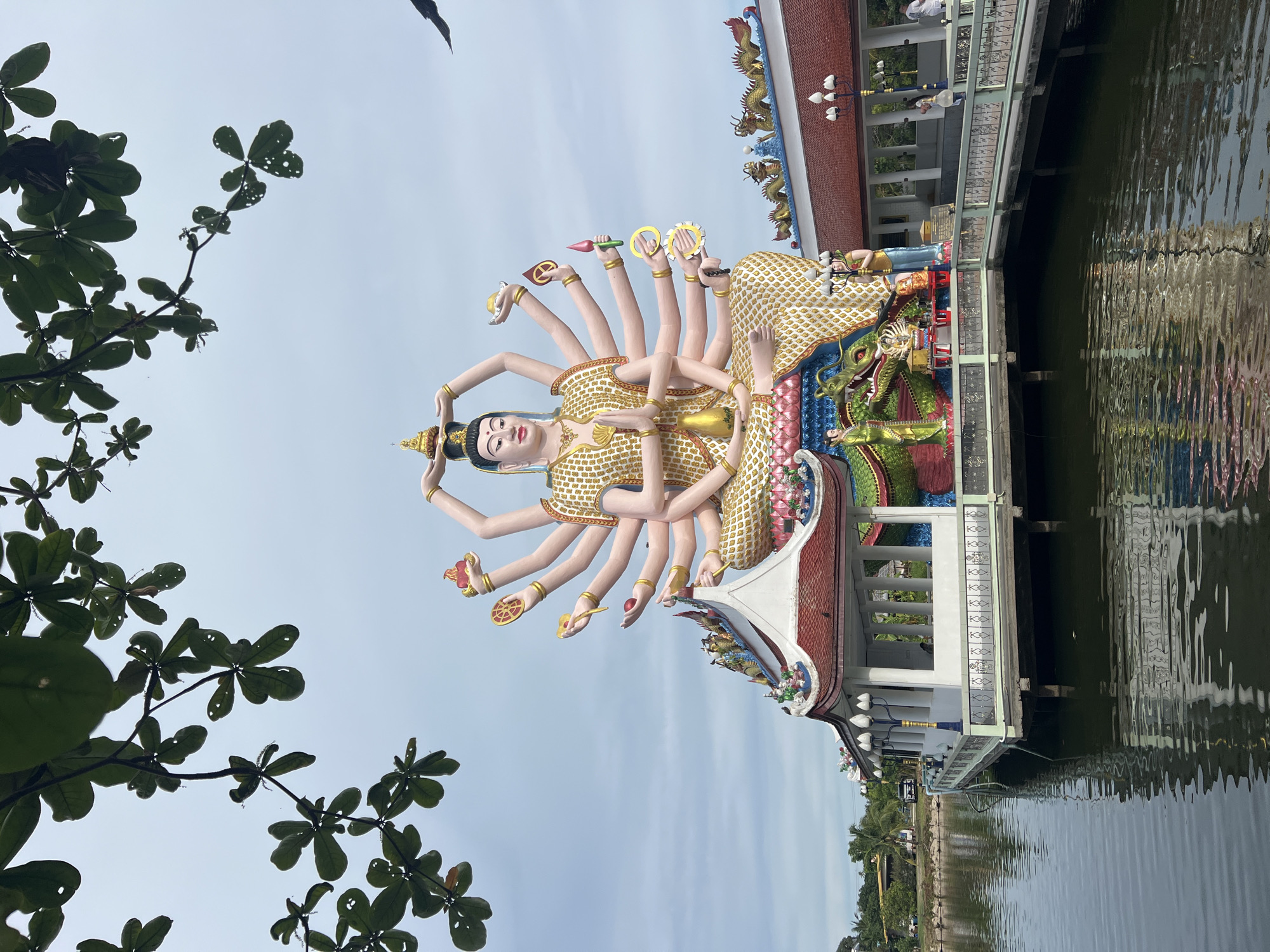 The image depicts the exterior of the Baan Plai Laem temple complex in Koh Samui, Thailand. Dominating the scene is a large, multi-armed statue of a deity, seated in a meditative pose atop a colorful structure that appears to be floating on a body of water. The statue wears an ornate golden robe and holds various symbolic objects in its many hands. The temple buildings feature traditional Thai architectural elements, including ornate roofs with dragon motifs. The water surrounding the temple reflects the structures and the overcast sky. Lush green foliage frames the top of the image, obscuring parts of the sky. A bird is visible in flight in the upper right portion of the frame. A few individuals, dressed in white, are seen walking on a walkway to the right of the main statue, suggesting the temple is open to visitors. The overall atmosphere appears serene and spiritual.