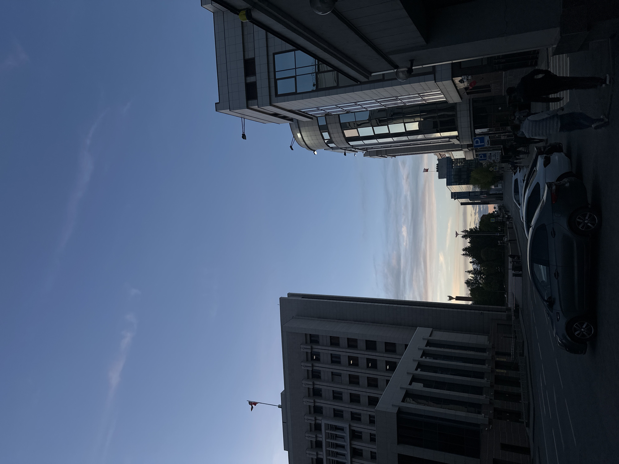 An eye-level, horizontal photograph looking up at modern buildings from the street in Kazan, Russia. The buildings are primarily white and grey, with a glass facade. One building includes a flag at its peak, along with windows. Other buildings include light fixtures and architectural detailing. Two people are seen walking on the sidewalk near parked cars. In between the buildings, there is a row of trees near the horizon. The sky is blue, with light clouds and visible sunlight.