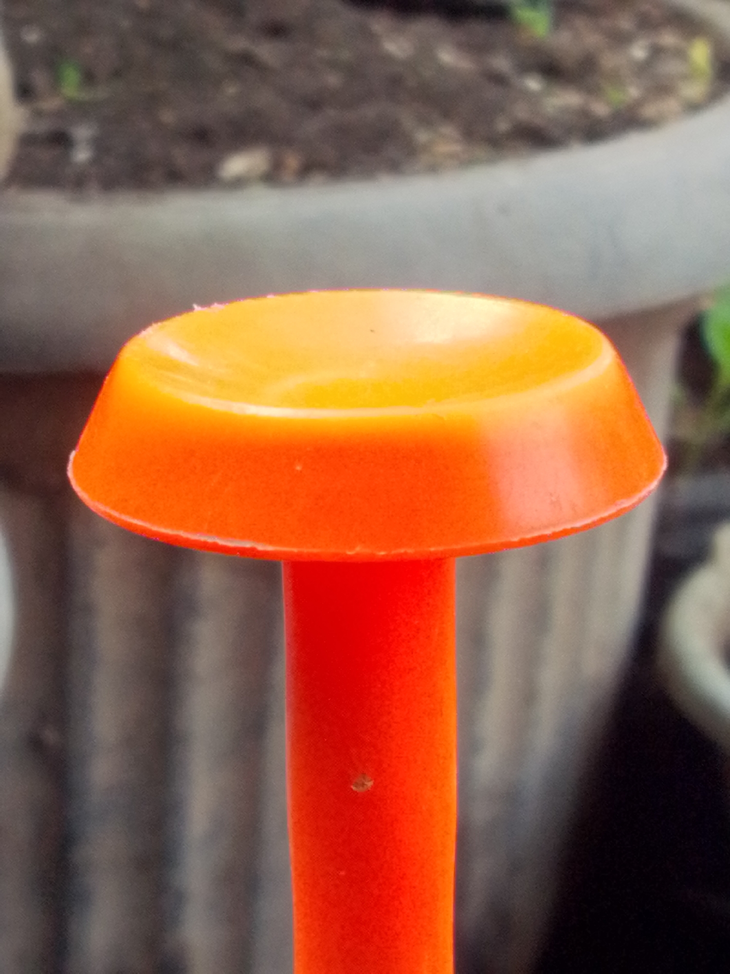 A close-up, eye-level shot focuses on the top of an orange plastic gardening tool, possibly a dibbler or planter, with a conical shape that flares out at the top. The surface is slightly textured and reflects light, showing a gradient from a brighter orange at the top to a deeper orange towards the bottom. The stem of the tool is a solid, bright orange cylinder. In the blurred background, the top of a large, grey, weathered pot is visible, filled with dark soil and hints of green foliage. The pot appears to be outdoors, with a textured, possibly concrete or stone surface. The lighting suggests it is daytime, with soft ambient light. The image does not depict any people or specific locations within Bandar Lampung, Indonesia, beyond a general outdoor garden setting.