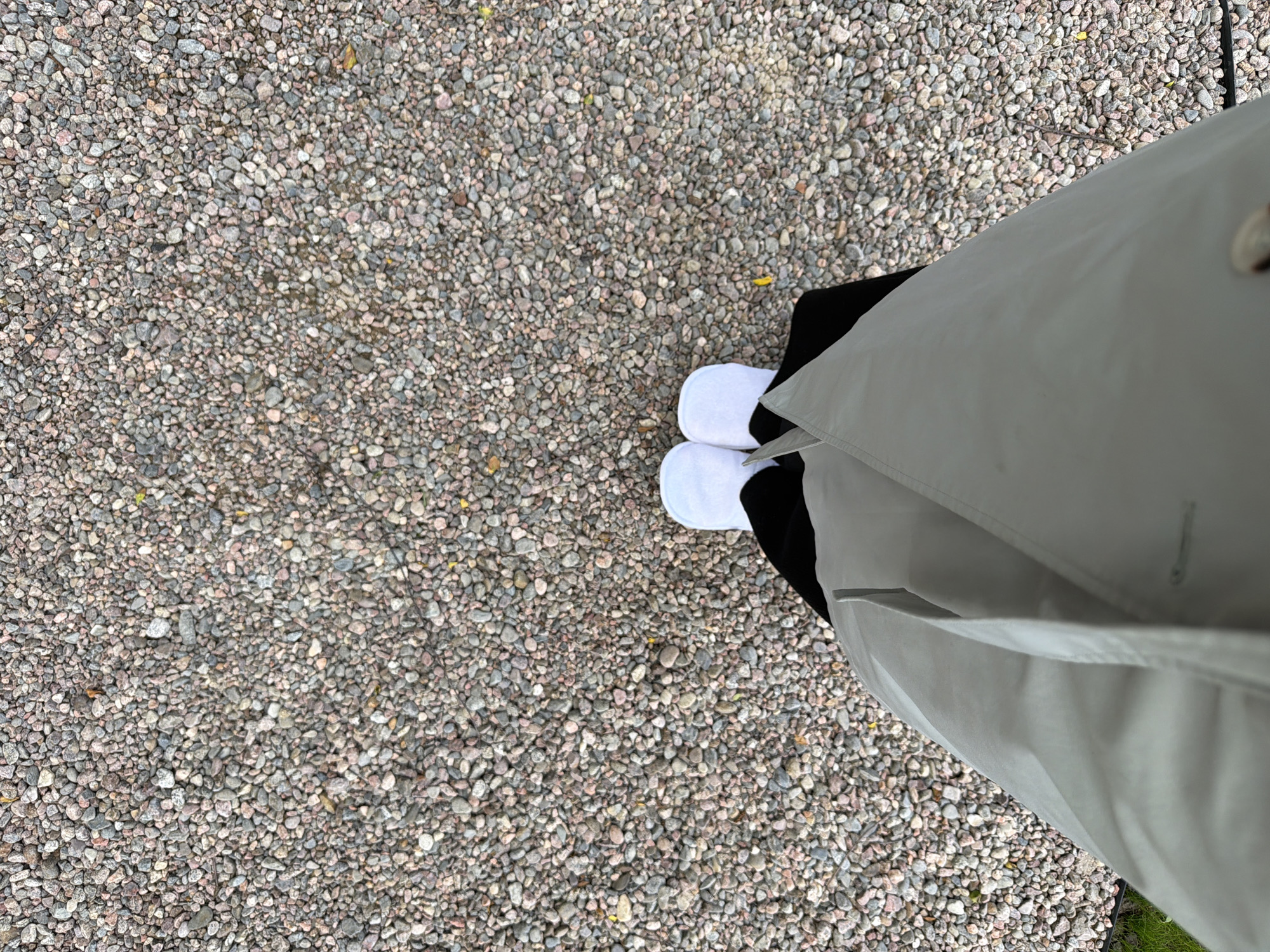 In Solov'evo, Russia, this image captures a person's feet clad in white socks and slippers, standing on a ground covered with small, varied-colored gravel. The person is wearing a light grey trench coat over black clothing. The scene is viewed from a high angle, looking down at the person's lower body and the surrounding gravel path.