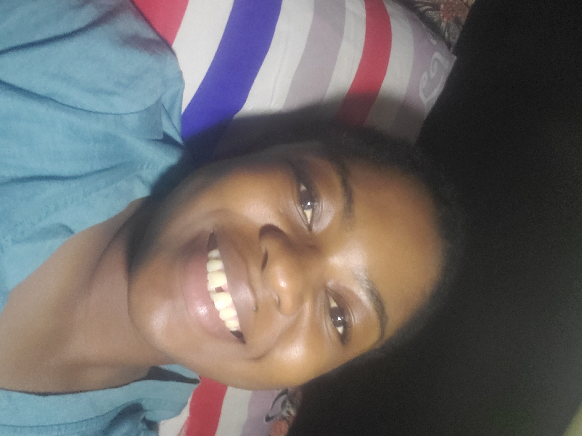 This image depicts an indoor scene in the city of Kakau, Nigeria. A dark-skinned adult female is centrally positioned, smiling broadly and looking directly into the camera. Her face exhibits a natural sheen. She has dark eyes, full lips, and dark, natural hair. Her upper teeth are visible, featuring a distinct gap between the two central incisors. She is wearing a light blue short-sleeved top. She appears to be reclining, with her head resting on a pillow adorned with horizontal red, white, and blue stripes, visible on the left side of the frame. A darker, patterned fabric is partially visible below the pillow. The background on the right and upper portions of the image is dark and indistinct.