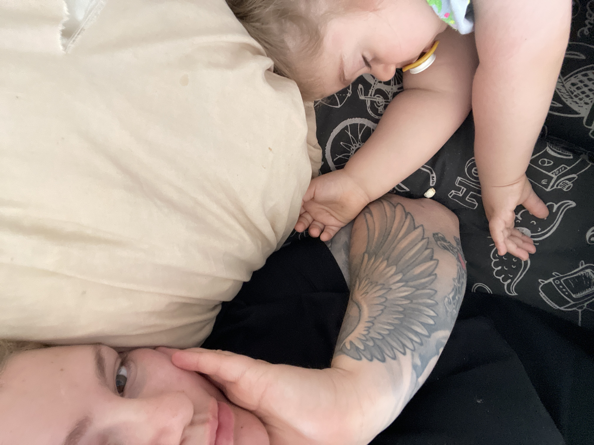 A woman lies in bed, possibly in Senden, Germany, based on location instructions, with a sleeping baby resting on a pillow near her. The baby has a pacifier in its mouth. The woman is looking at the camera, holding her face with her hand. She has a wing tattoo on her arm. The baby is lying on a black and white patterned sheet.