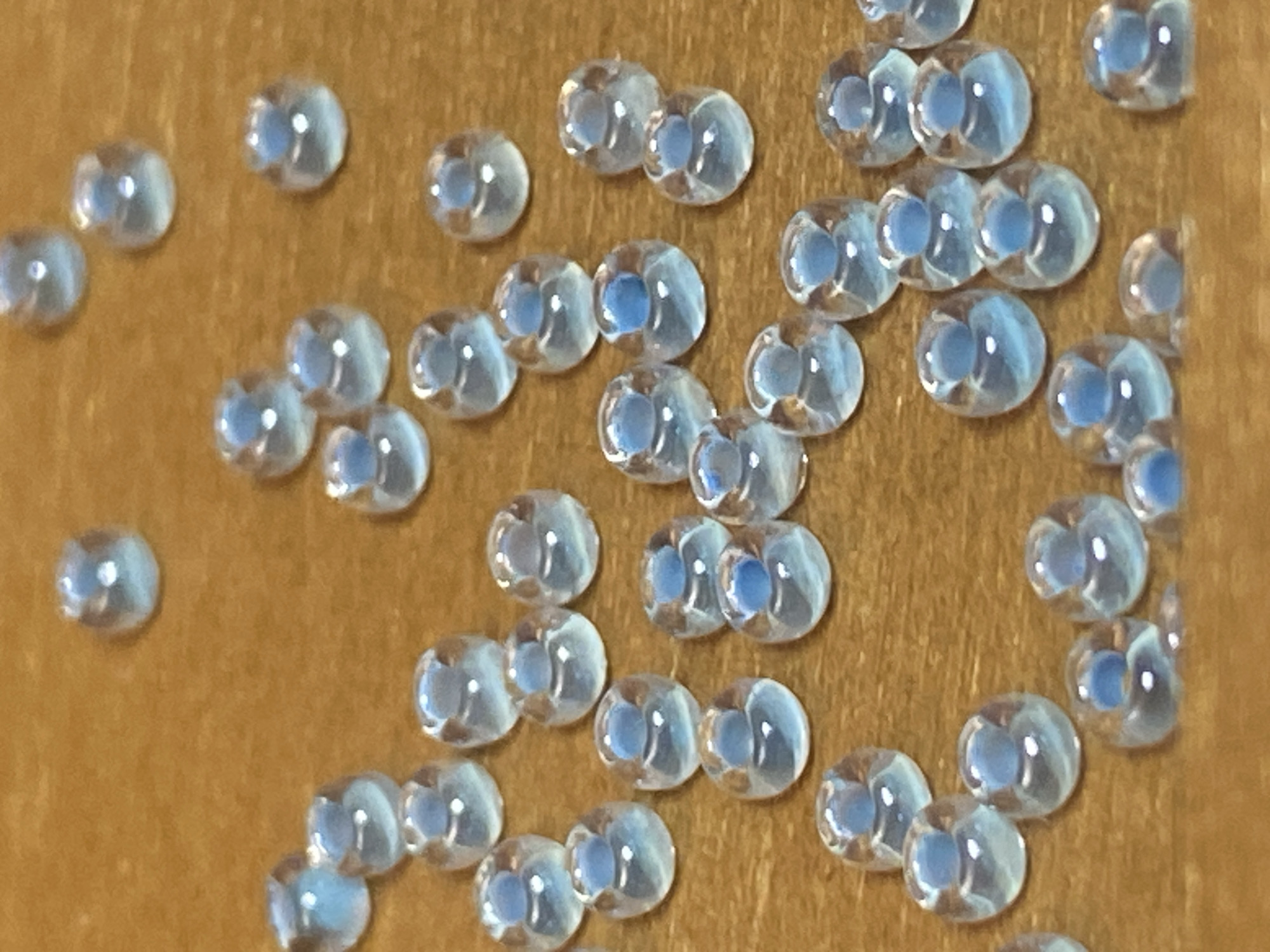 The image shows a close-up, overhead view of numerous small, translucent, light blue beads scattered on a warm brown, wooden surface. The beads are oval-shaped with a hole through the center, and they appear to be made of glass or a similar transparent material that reflects light. Some beads are stacked or slightly overlapping. The lighting appears to be artificial, creating highlights and shadows on the beads and the wooden surface. There are no people, animals, or other objects visible in the frame. The context of Saint Petersburg, Russia is not discernible from the visual information in the image.