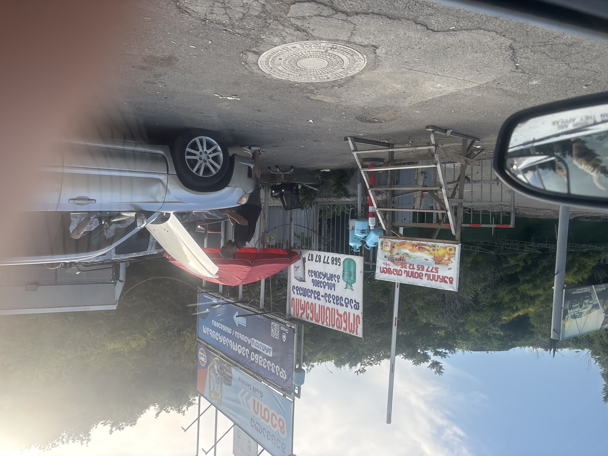 The image depicts an outdoor scene, likely a roadside service area in Tbilisi, Georgia, bathed in bright sunlight. In the foreground, a silver SUV with its hood open is being attended to by a man. The SUV has a dent on its passenger side front fender. Behind the SUV, a red umbrella provides shade over a work area, and a swivel chair is visible. To the left, several blue propane tanks are stacked near a wooden structure that resembles scaffolding or a ladder.
Several signs are present. One large billboard in the background advertises "Gulf Service" with an image of car oil. Another sign, in Georgian script, appears to be for a gas station or service point, listing a phone number "577 77 87 07" and mentioning propane. A smaller sign to the left also features Georgian script and an image of honey, with the phone number "577 65 12 35". A striped gate barrier is visible in the middle ground.
The ground is asphalt, with some cracks and debris. A manhole cover with intricate detailing is prominent in the lower center of the frame. In the reflection of a car's side mirror on the left, a cat can be seen. The background is dominated by lush green trees and foliage on a hillside, under a bright, partly cloudy sky. The overall impression is a functioning, albeit somewhat cluttered, service stop.