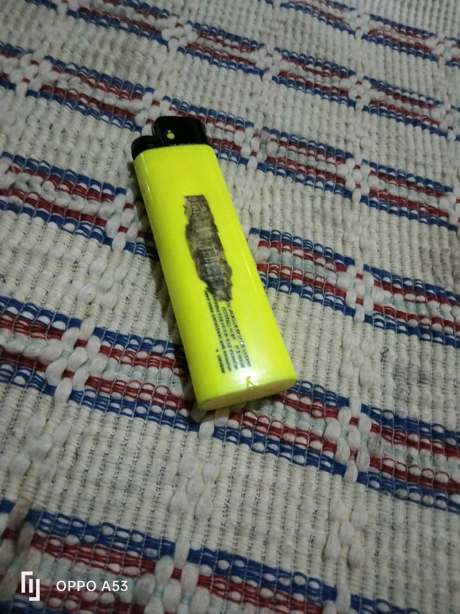 A bright yellow lighter with a black top rests on a textured, woven surface. The surface displays a repeating pattern of horizontal stripes in white, blue, and red. The lighter has a dark, smudged mark on its body, possibly from use. Text is visible on the lighter's surface, but it is too blurred to read. The bottom left corner of the image shows the text "OPPO A53," indicating the device used to capture the photograph. The overall scene is an indoor shot, likely taken in a home environment in Trenggalek, Indonesia, given the visible device model often associated with that region.