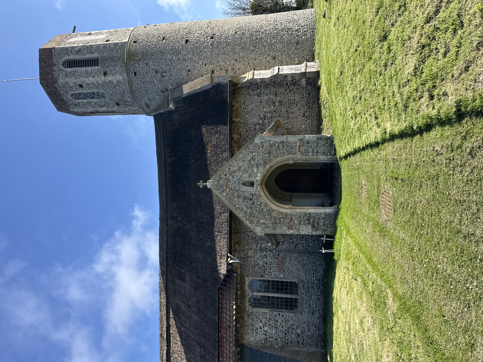 This media file presents a historical stone church, typical of the East Anglian region in the United Kingdom, consistent with the provided location context of Great Yarmouth. The image is taken from a low angle, looking upwards at the church against a bright blue sky scattered with wispy white clouds.
The most prominent feature is the church's distinct round tower, constructed from textured, light-colored flint or local stone. The tower transitions from a cylindrical lower section to an octagonal belfry at its summit, which includes arched, louvered openings. A flagpole with a finial extends from the very top of the tower. The main body of the church features a dark, possibly thatched or shingled roof, while other sections exhibit terracotta tiling.
An arched stone porch, also built from flint, serves as the main entrance to the church, topped with a small cross on its gable. A leaded glass window with a stone frame is visible on the church's side wall. The church is situated within a well-maintained green grassy area, likely a churchyard, where a faint, worn path leads towards the entrance. Two small, white crosses are subtly visible in the grass, indicating graves or memorials.
The scene is captured in bright daylight, with clear shadows on the grass suggesting either mid-morning or afternoon. No people or specific activities are visible, lending the image a serene and timeless quality. There is no visible text in the image.