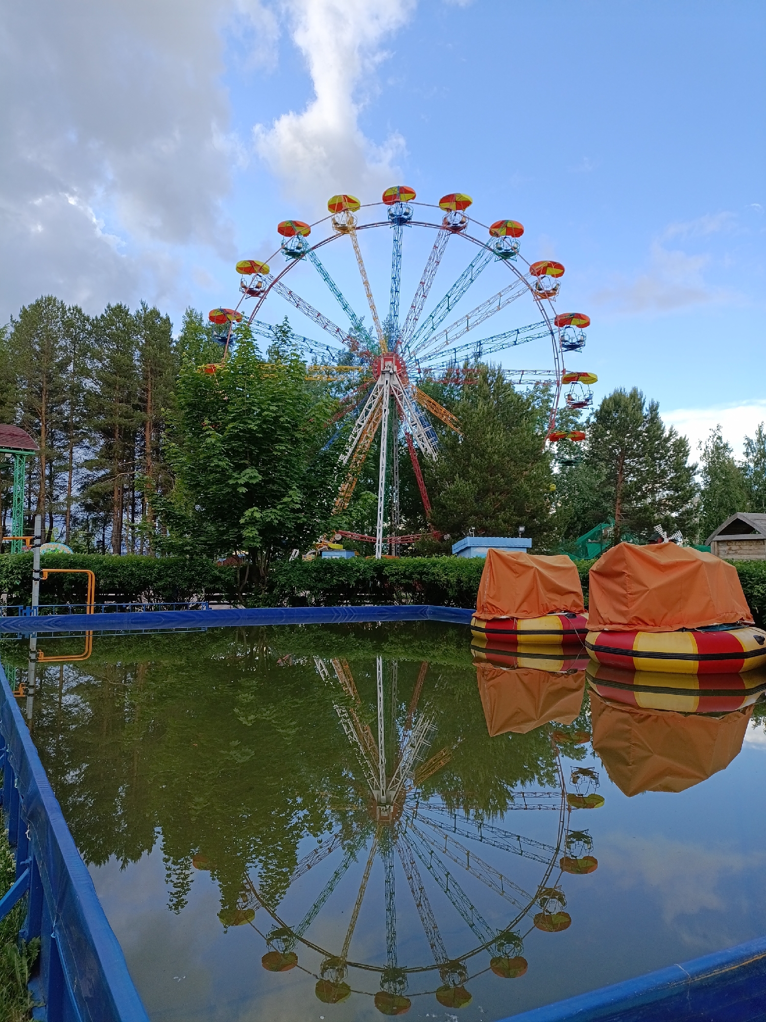 The image depicts a quiet amusement park scene in Chaikovsky, Russia, under a partially cloudy sky. A large, colorful Ferris wheel dominates the background, its intricate metal structure and cabins vividly reflected in a rectangular pool of water that occupies the foreground. Its cabins display red, yellow, blue, and green accents, and its nearly perfect reflection shimmers on the pool's still, somewhat murky surface.
In the immediate foreground, two bumper boats, covered with orange tarps over their red, yellow, and black striped bases, float idly in the pool. A blue fence borders the left side of the pool. Lush green trees, including tall pines and deciduous varieties, create a dense backdrop behind the pool and around the Ferris wheel, suggesting a natural park environment. Subtle hints of other amusement park structures are visible through the foliage.
The sky features a mix of clear blue patches and soft, greyish-white clouds. The absence of people lends a quiet, contemplative atmosphere; the covered boats suggest they are not in use, perhaps out of season or after hours. Soft lighting, devoid of harsh shadows, hints at late afternoon or early evening. No visible text is present in the image.