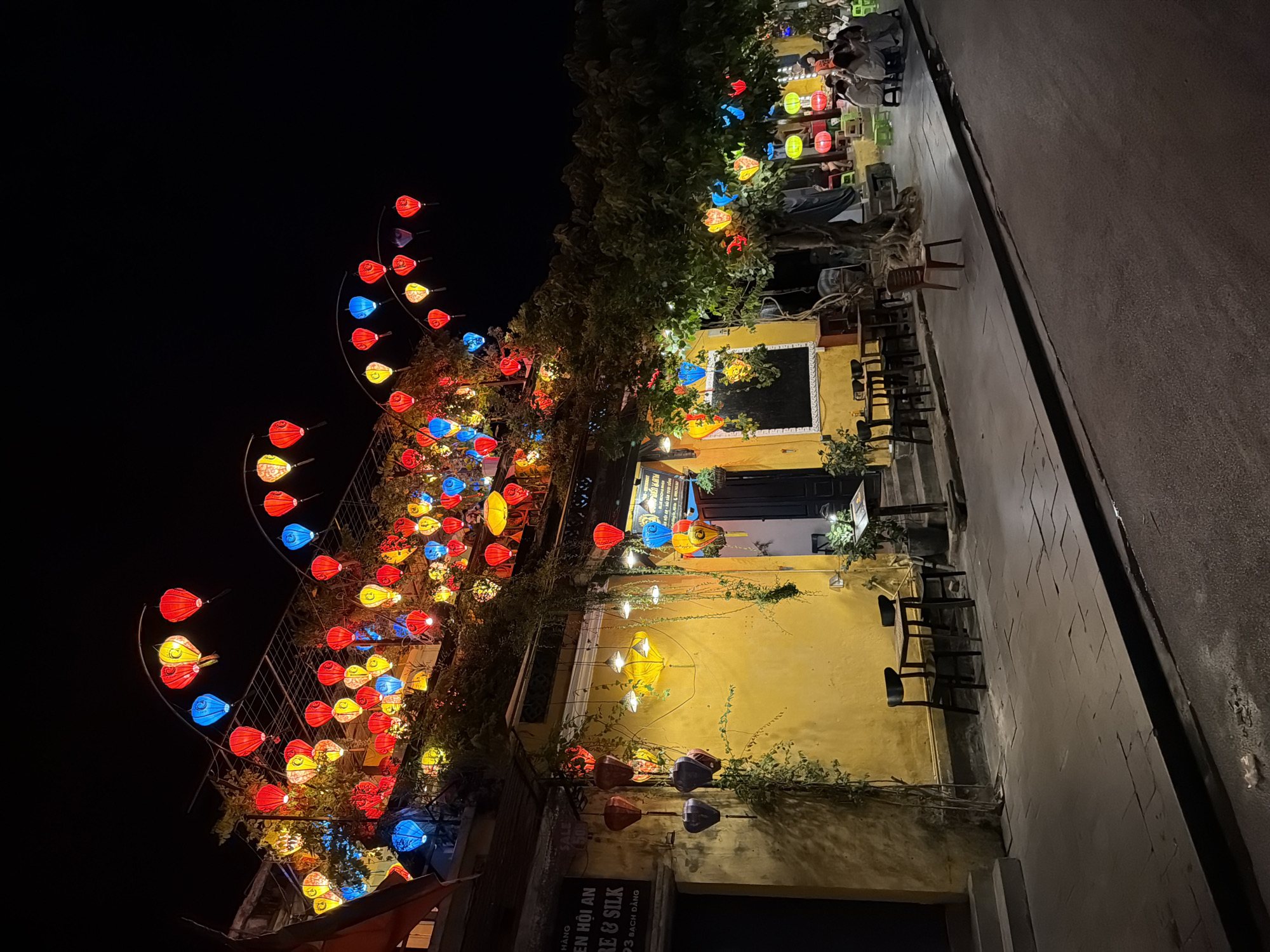 The image depicts a street scene at night in Hội An Ward, Vietnam, characterized by vibrant, illuminated lanterns. The foreground shows a paved sidewalk adjacent to a street. On the sidewalk, there are several outdoor seating arrangements with tables and chairs, some occupied by patrons. A large tree with branches adorned with colorful lanterns stands prominently in the midground. The background features a building with a bright yellow facade, also decorated with numerous lanterns hanging from its exterior and roofline. A sign with Vietnamese text is visible on the building. The overall atmosphere is festive and atmospheric, illuminated by the glow of the lanterns against the dark night sky.