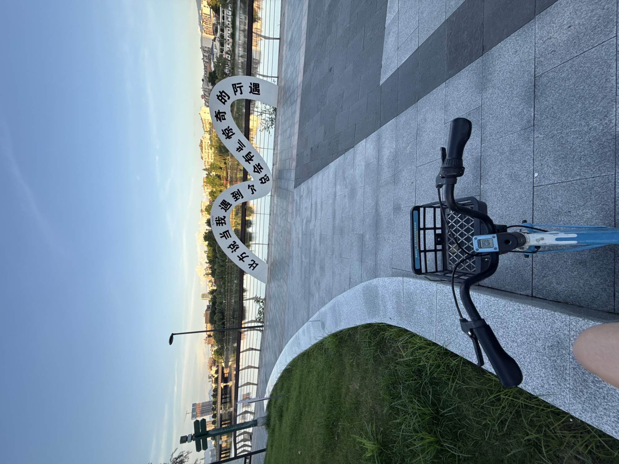 An outdoor shot captures a shared bicycle parked on a paved walkway in Yiwu, China, during daylight. The bicycle, primarily blue and white, faces forward with its handlebars and basket prominently visible. To the left of the bicycle, a curved patch of green grass borders the walkway, transitioning into a wider paved area. In the background, a river reflects the sky and the cityscape, which includes various buildings and trees. A distinctive white, wavy sculpture with black Chinese characters is situated near the river, adding a cultural element to the scene. The sky is clear and blue, with a hint of clouds. The overall environment appears to be a park or waterfront area within the city.