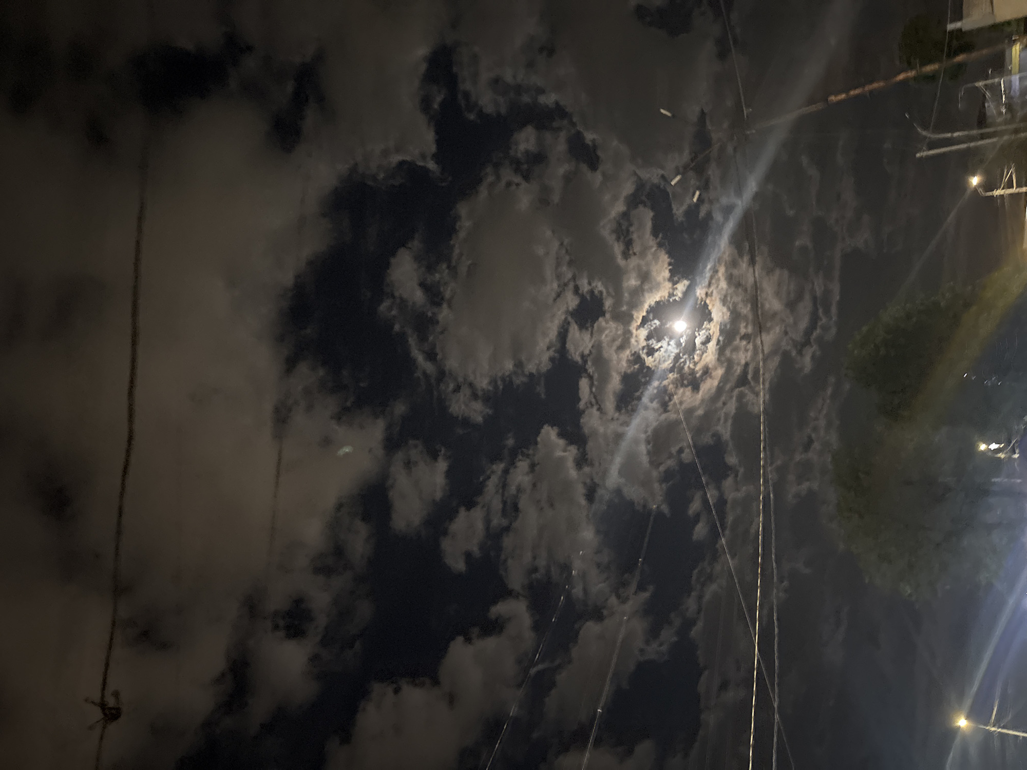 This is a low-angle, nighttime photograph depicting a cloudy sky illuminated by a bright light source, likely the moon. Several dark electrical wires intersect the sky. In the upper right corner, there's a blurred view of buildings and trees, indicating an urban location. The picture has elements of Yerevan's urban infrastructure and atmospheric conditions.
