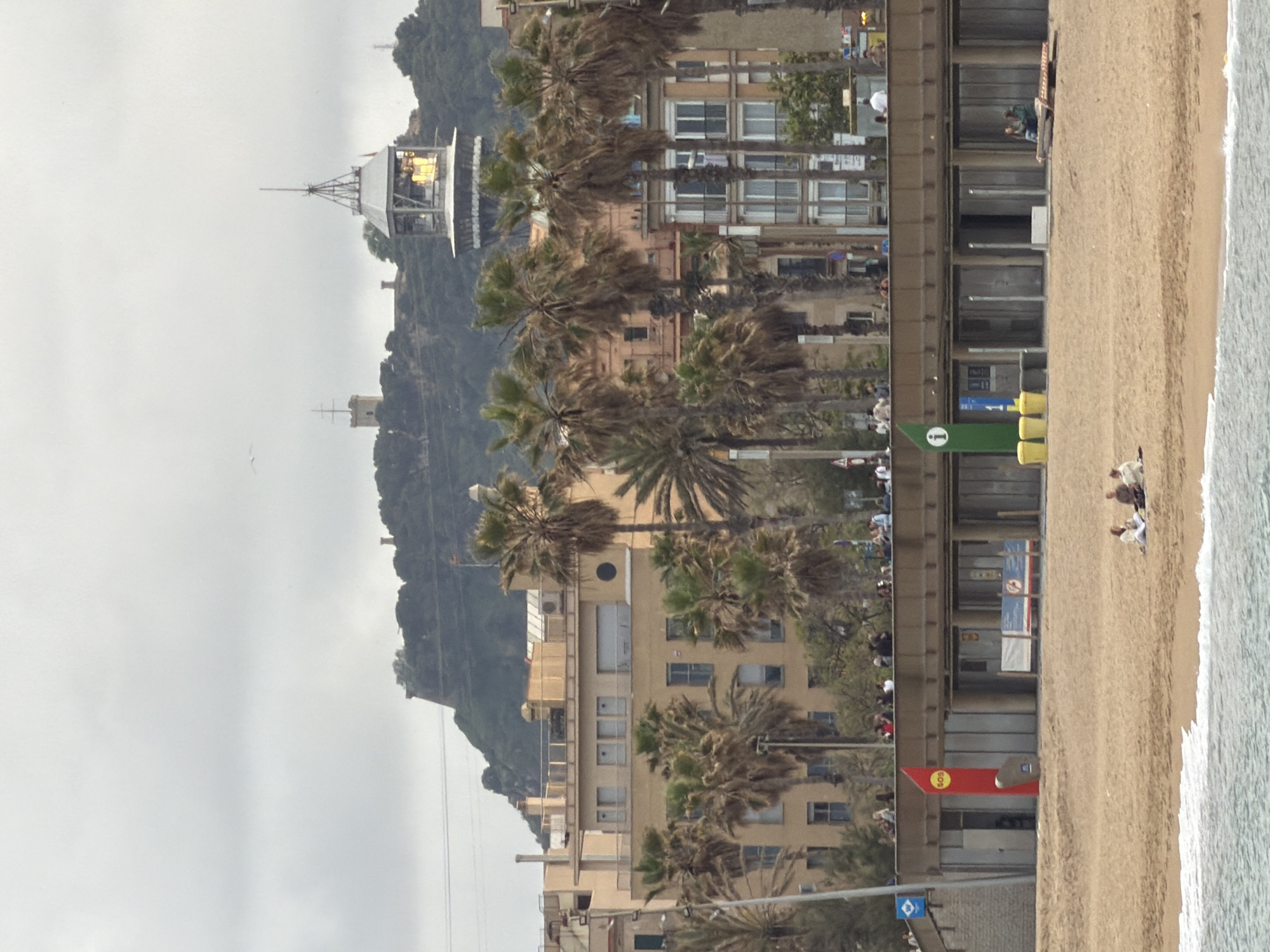 The image shows a coastline scene in Barcelona, Spain. From left to right, a hazy sky dominates the upper portion, with a mountain range beneath. A structure resembling a cable car station sits atop the mountain, and buildings with palm trees line the shore, separating the mountain from the beach. On the beach, people are visible, with some appearing to be sitting near the water's edge. The right side of the image shows sand leading to the ocean's edge. Signage and structures along the beach indicate a public access area.