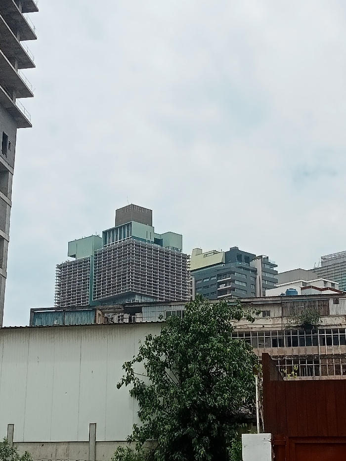 This image captures an urban landscape in Luanda, Angola, on an overcast day. The scene is dominated by a mix of modern and older buildings under a uniformly grey, cloudy sky.
In the left foreground, a tall, partially constructed concrete building with exposed floor slabs and rebar rises vertically, indicating ongoing development. To its right, in the mid-ground, stands a distinctive, multi-story building featuring a prominent gridded facade, possibly a Brutalist or mid-century modern design. The upper sections of this building incorporate lighter green or teal-colored panels and a darker, box-like structure on top.
Further to the right and in the background, several other modern buildings are visible, some with dark grey or black facades, balconies, and accents of the same green/teal color seen on the central building. Below these, a long, low-rise structure stretches across the mid-ground, characterized by a corrugated metal roof and numerous windows or openings along its facade. The left portion of this lower structure presents a plain, light-colored wall.
A lush, leafy green tree occupies the lower mid-ground, partially obscuring the base of the lower buildings. In the far lower right, a dark brown, possibly metallic, fence or gate is visible.
There are no people or specific activities taking place, and no discernible text is visible. The time of day appears to be daytime, though the heavy cloud cover prevents any direct sunlight, contributing to a muted and somewhat industrial atmosphere.