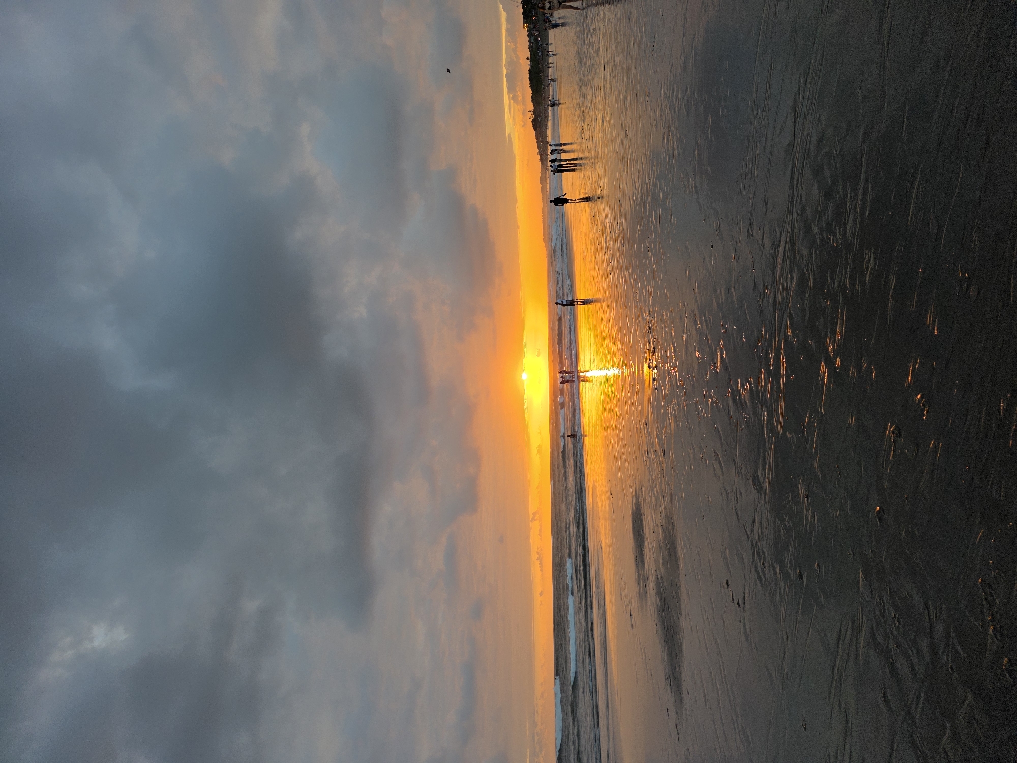 The image depicts a sunset scene at Seminyak Beach in North Kuta, Indonesia. The sky is filled with dramatic, dark gray clouds that are partially illuminated by the setting sun. The sun, a vibrant orange orb, is low on the horizon, casting a warm glow across the water and the wet sand. The ocean is calm, with gentle waves rolling towards the shore. Several people are visible on the beach, some walking, some standing by the water's edge, and a few appear to be interacting with each other. The wet sand reflects the colors of the sunset, creating a shimmering, golden path. The overall atmosphere is serene and picturesque, capturing the beauty of a tropical beach at dusk.