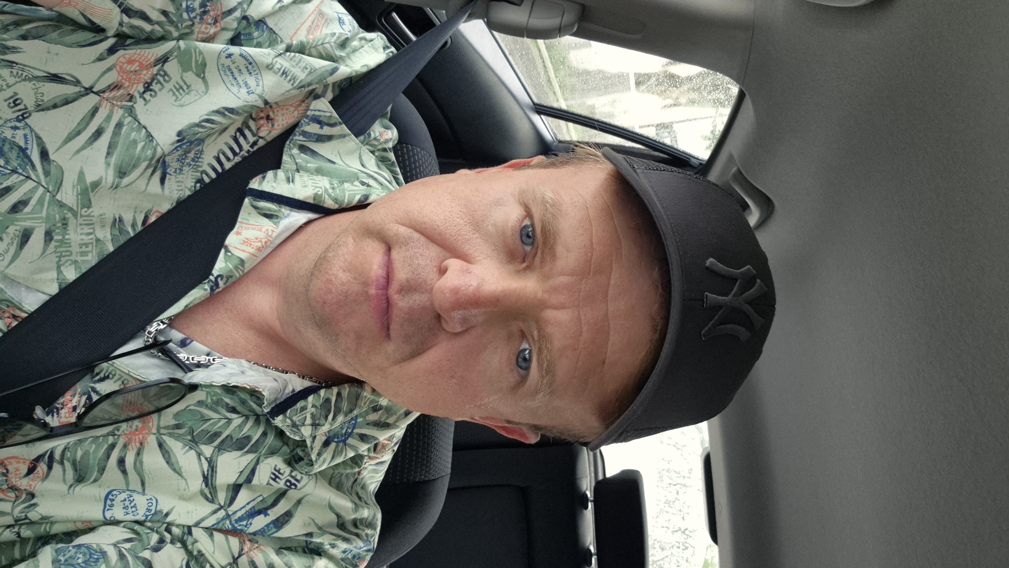 A middle-aged man with a baseball cap is sitting in a car, wearing a floral shirt and a seatbelt. The man is fair-skinned with blue eyes and light-colored hair. The baseball cap has the "NY" logo on it. A pair of glasses are hanging from the collar of the shirt. The photo appears to have been taken from a slightly elevated angle and has been rotated counter-clockwise. The background shows a car interior and a glimpse of a rainy landscape through the window. There is no evidence in the image to suggest the location in Ergolding, Germany.