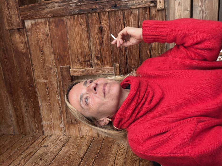 A solo adult female is depicted in a medium shot, seated in a wooden structure in La Clusaz, France. She has blonde hair and wears a bright red cowl-neck sweater with ribbed cuffs and gold-toned hoop earrings. Her head is tilted slightly upwards, and her gaze is directed towards the upper left, exhibiting a neutral expression. Her right hand is raised, holding a lit white cigarette with a visible filter between her index and middle fingers. The background features aged, horizontally-oriented wooden plank walls, characterized by natural grain and knots. To the right of the subject's head, a small, dark rectangular opening, framed by vertical wooden beams, is integrated into the wooden paneling.