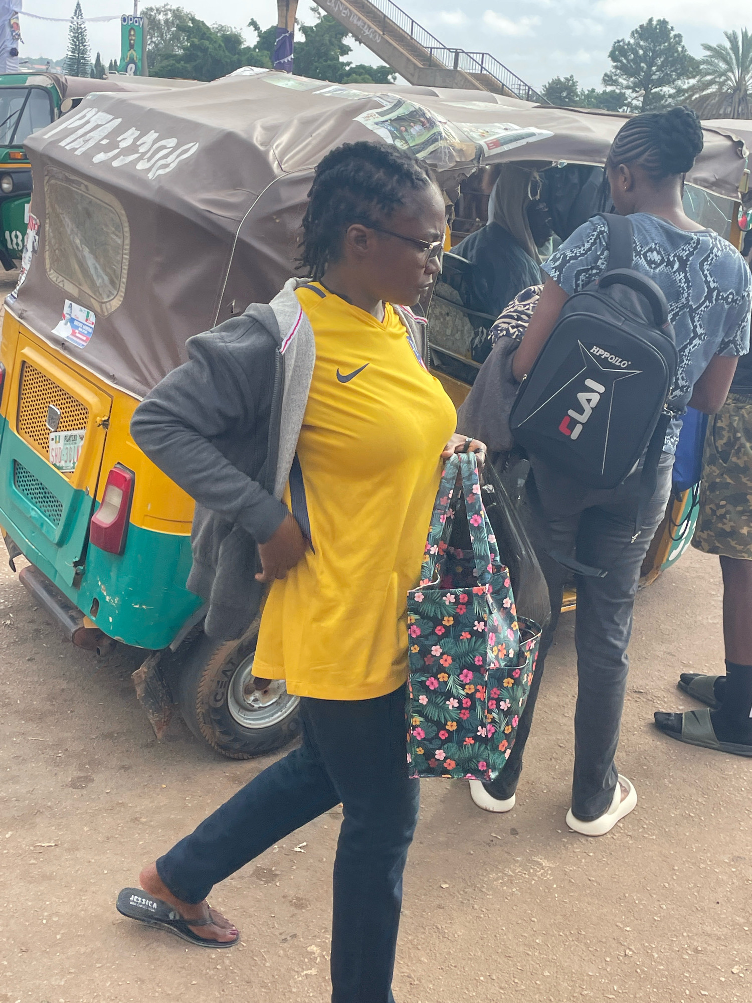 The image depicts a street scene in Gura Top, Nigeria, with a focus on several individuals near a colorful auto-rickshaw. In the foreground, a woman wearing a yellow t-shirt and a grey jacket is walking, carrying a floral patterned tote bag. She is wearing dark pants and black flip-flops with the brand name "Jessica" visible. To her right, another woman is standing, wearing a blue and black patterned t-shirt and black pants, with a black backpack on her back that has "HPPOILO" and "ELAI" written on it. A third person, partially obscured, is visible behind them, wearing camouflage shorts and green slides. The auto-rickshaw, a common mode of transport in Nigeria, is parked to the left, exhibiting a yellow and green color scheme and a brown tarp covering its top. The ground is dusty and unpaved, indicative of a rural or less developed urban area. The background shows trees and a pedestrian overpass. The sky is overcast, suggesting a cloudy day.