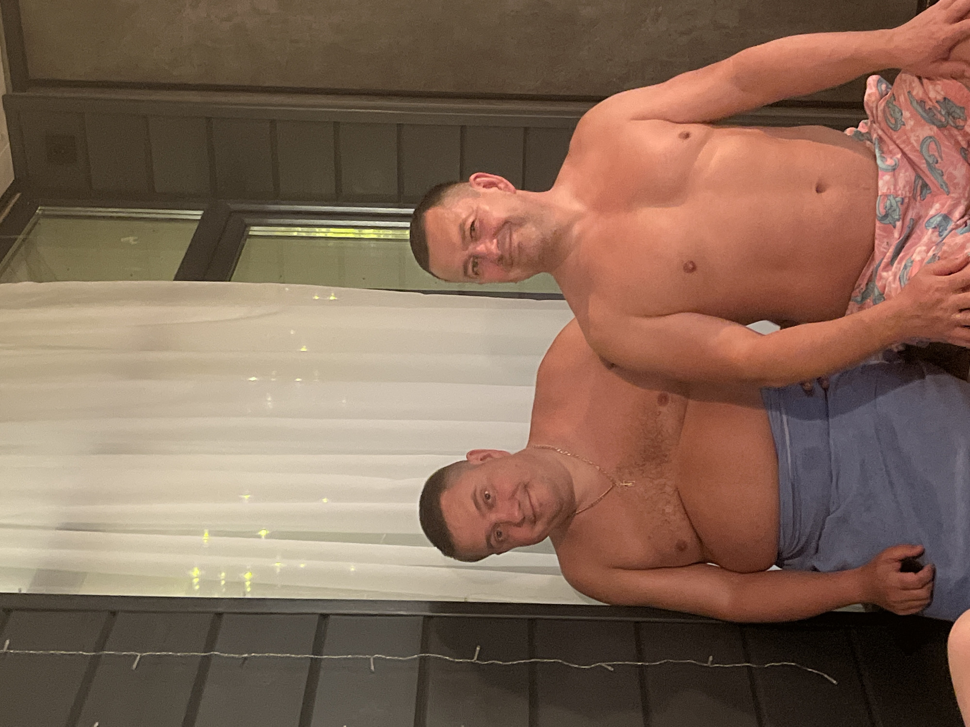 Two shirtless men are posing for a photo in what appears to be an outdoor setting. The man on top is wearing pink patterned shorts, while the man below is wearing blue shorts. The location includes a window and a white curtain. The background also has some string lights. This appears to be a candid, informal photograph. There is no specific information linking the image to the city of Новая Чернушка, Russia.